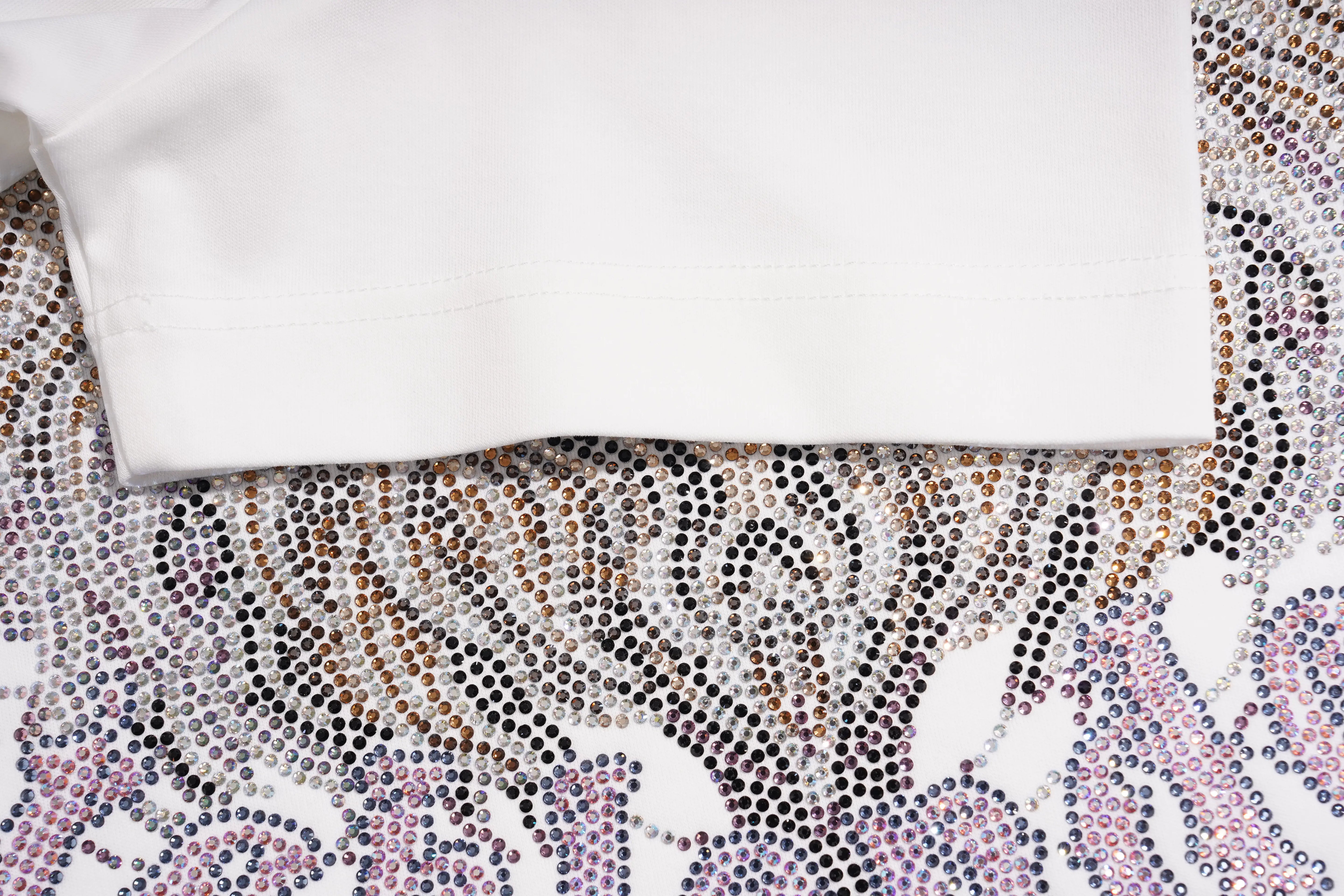 A close-up of a white folded garment laid on top of a heavily embellished patterned fabric featuring rows of rhinestones in black, gold, brown, and pink. TCH Regal Crown Lion Rhinestone Graphic T-Shirt