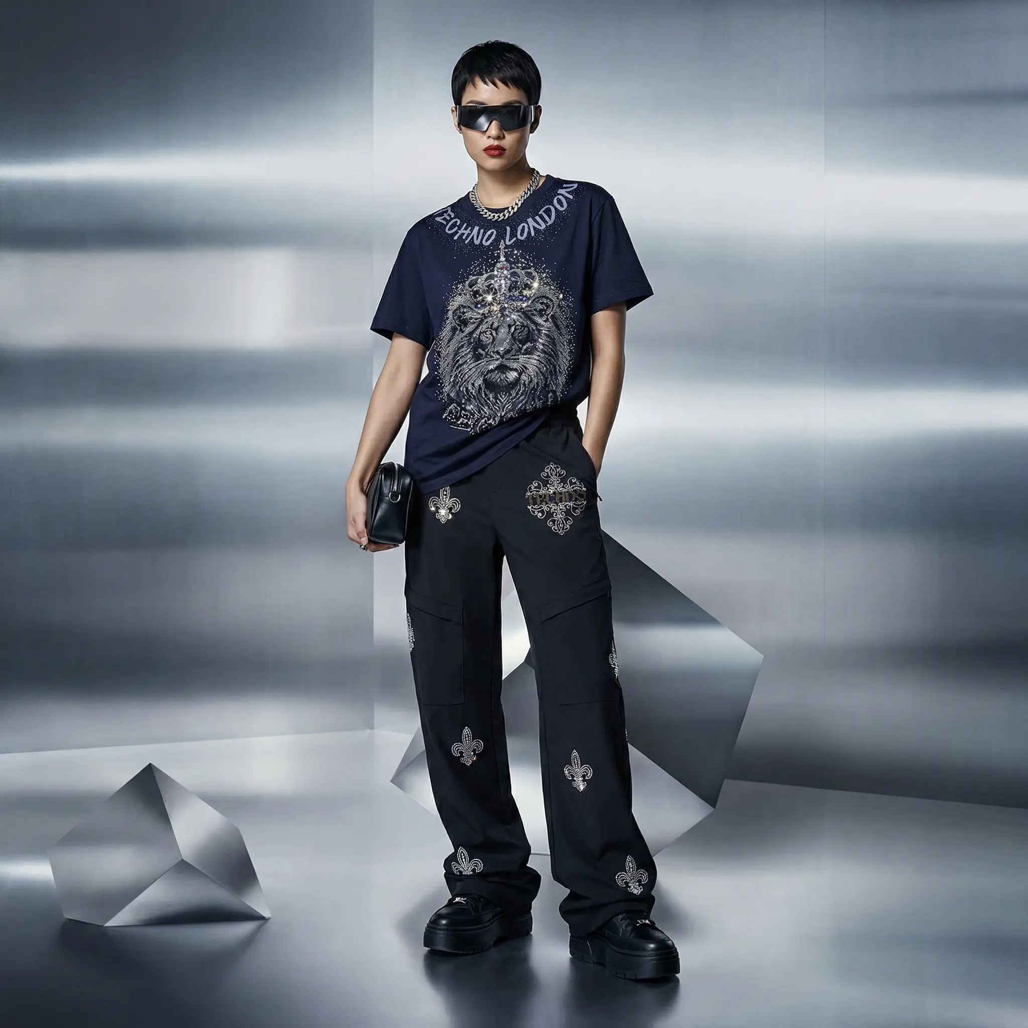 A model stands in a studio setting wearing a dark navy graphic T-shirt with a large lion print and 'SEMPER LONDON' text, paired with matching dark pants featuring silver floral motifs. The background is a metallic silver-gray studio environment with geometric props. TCH Regal Lion Rhinestone Graphic T-Shirt