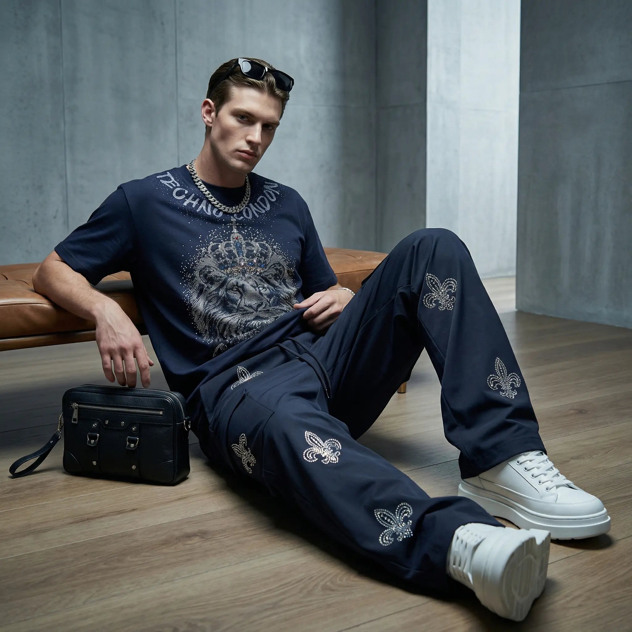 A man sits on a bench in a modern room wearing a navy blue graphic t-shirt and matching pants with white embroidered details, paired with white sneakers and a black crossbody bag. TCH Regal Lion Rhinestone Graphic T-Shirt