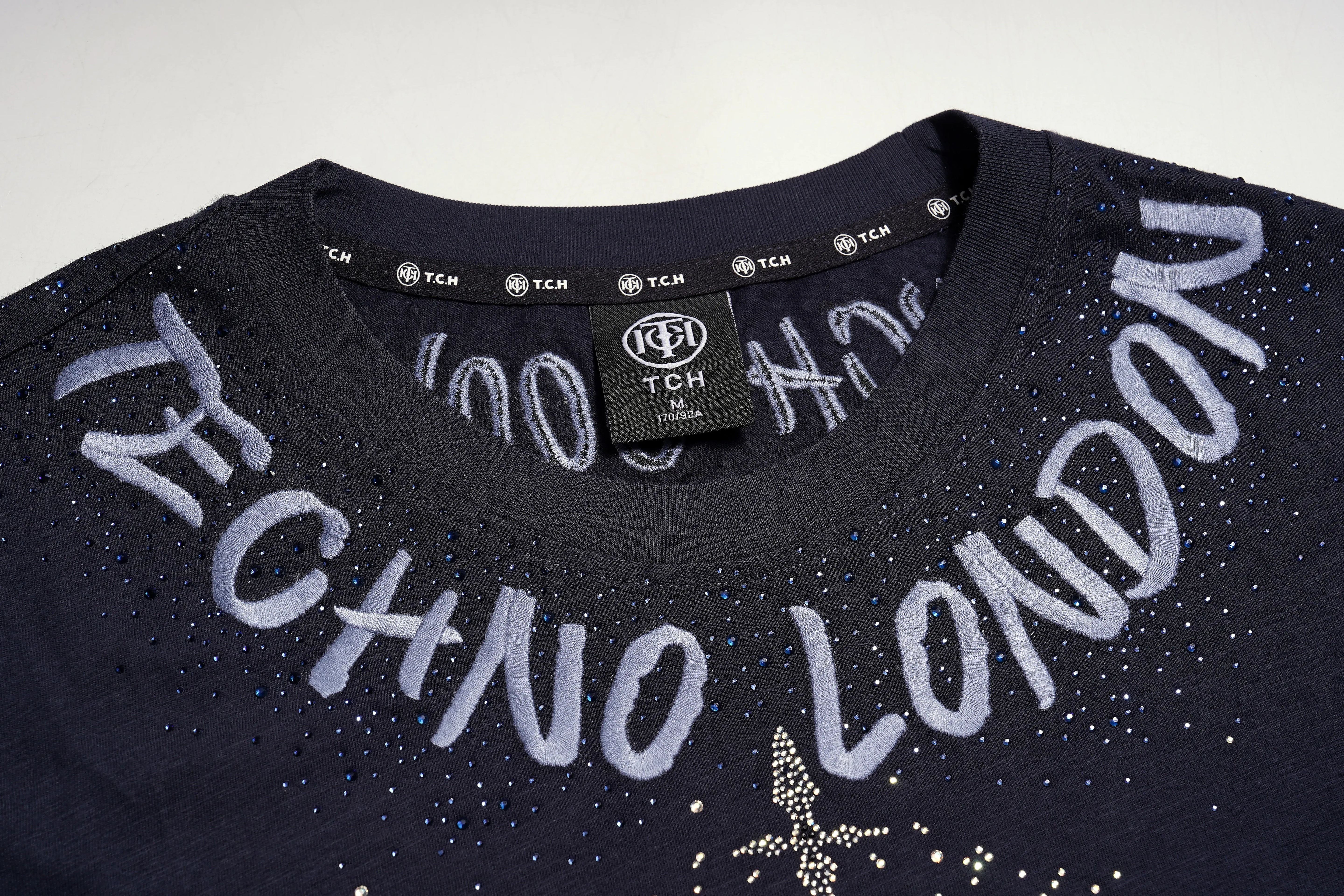 Close-up of a dark navy TCH London t-shirt featuring 'TECHNO LONDON' written across the chest in white rhinestone lettering, with a matching neck label and TCH branding visible. TCH Regal Lion Rhinestone Graphic T-Shirt