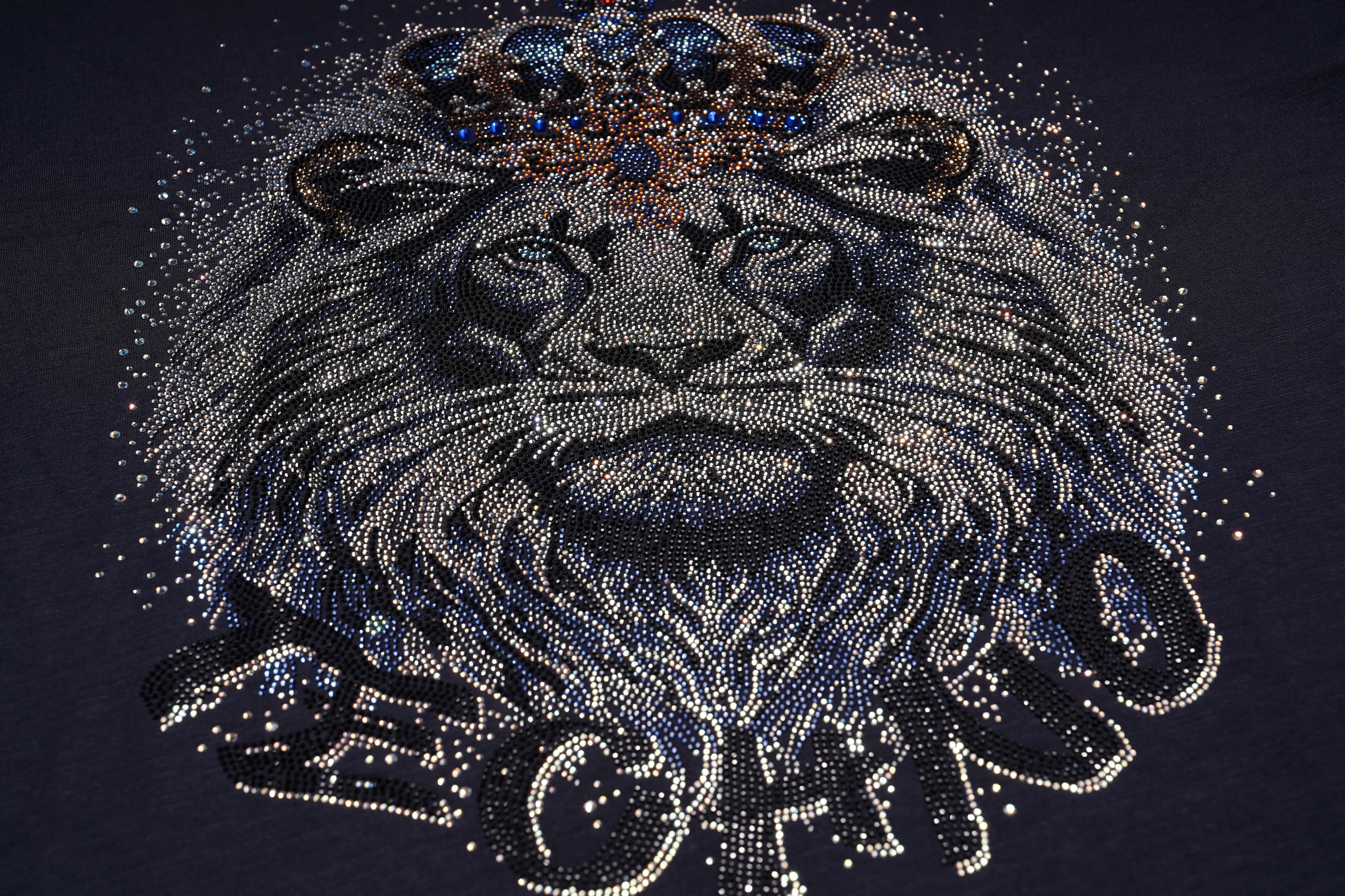 A close-up of a dark navy or black fabric featuring a large, ornate rhinestone lion graphic with a jeweled crown and stylized lettering below. TCH Regal Lion Rhinestone Graphic T-Shirt