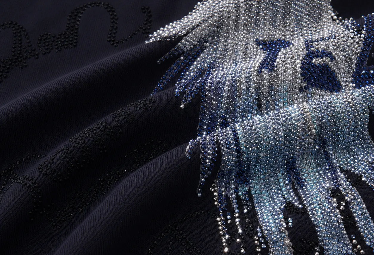 Close-up of a dark navy or black fabric with dense beaded embroidery and a large feather-like embellishment in silver, blue, and white tones. TCH Rhinestone Angel Wing Gradient Cargo Shorts