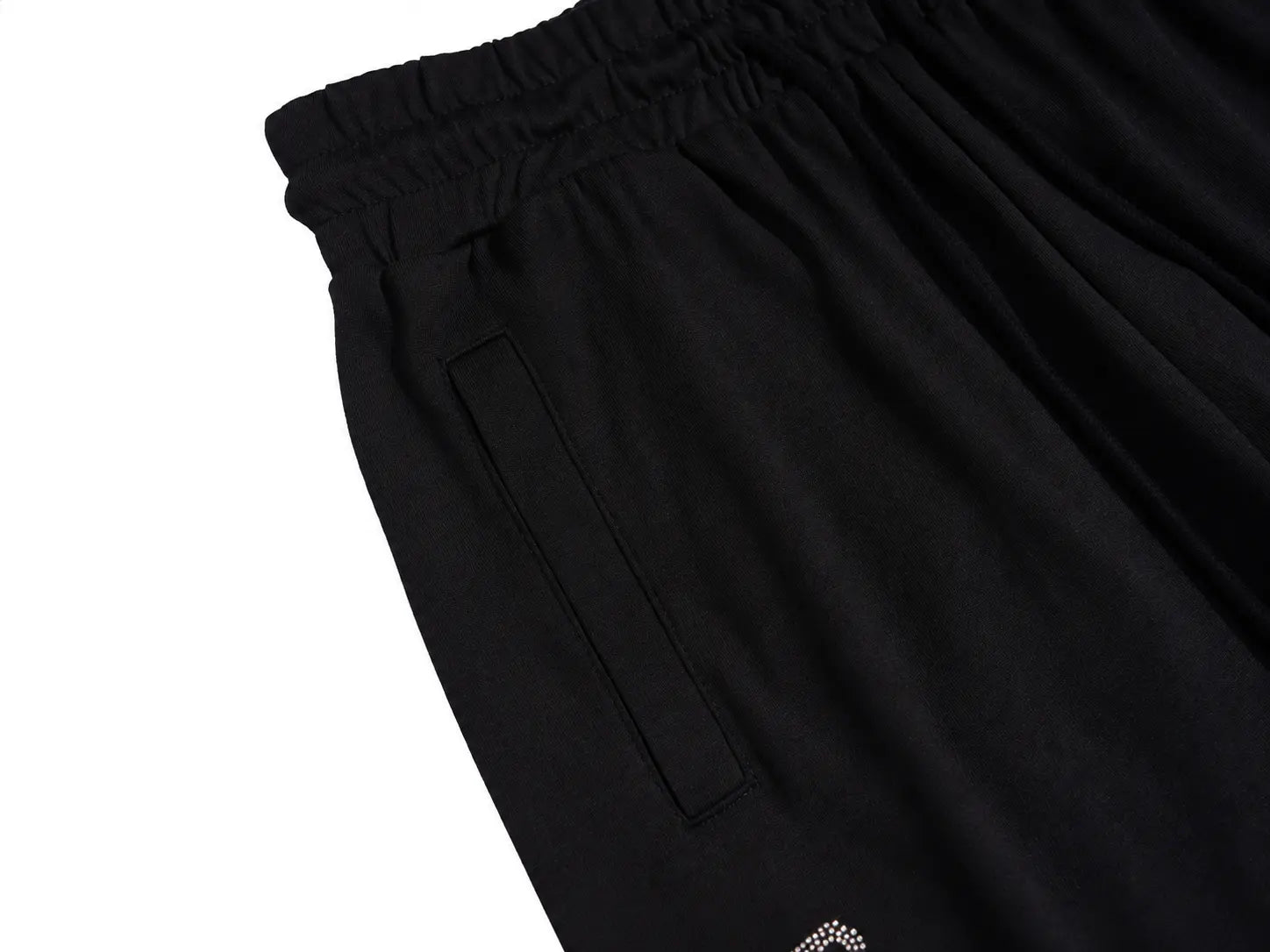 Close-up of black athletic shorts showing the elastic waistband, side pocket seam, and a small white embroidered logo near the lower hem, photographed on a white background. TCH Rhinestone Angel Wing Graphic Shorts