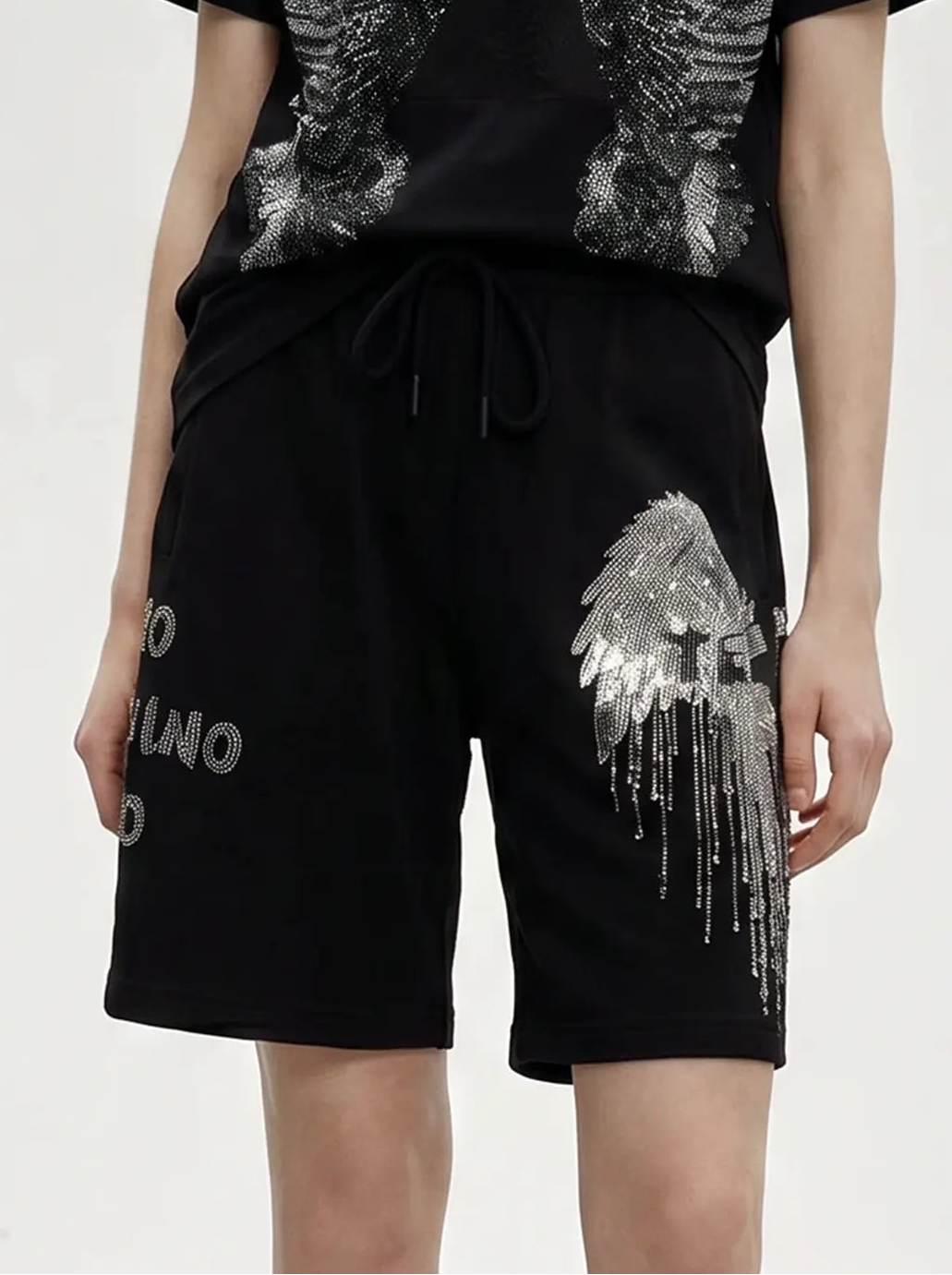 A model wearing a black short-sleeve t-shirt and matching black shorts, both featuring large distressed gray wing graphics and text details, set against a plain light gray studio background. TCH Rhinestone Angel Wing Graphic Shorts
