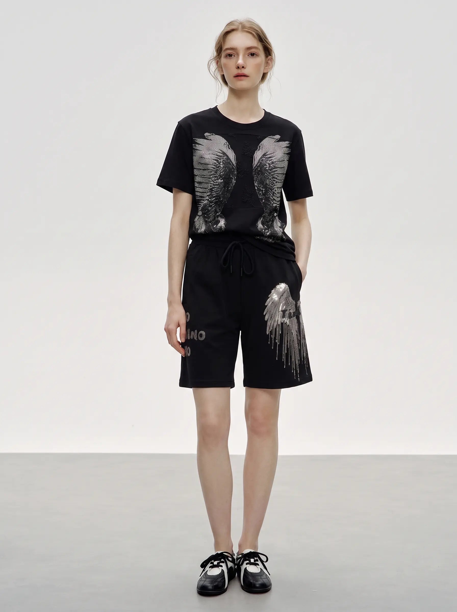 A model stands against a light gray studio background wearing a black short-sleeve T-shirt with large gray wing graphics on the front, matching black shorts with a gray wing graphic on the left leg, and black-and-white sneakers. TCH Rhinestone Angel Wing Graphic Shorts
