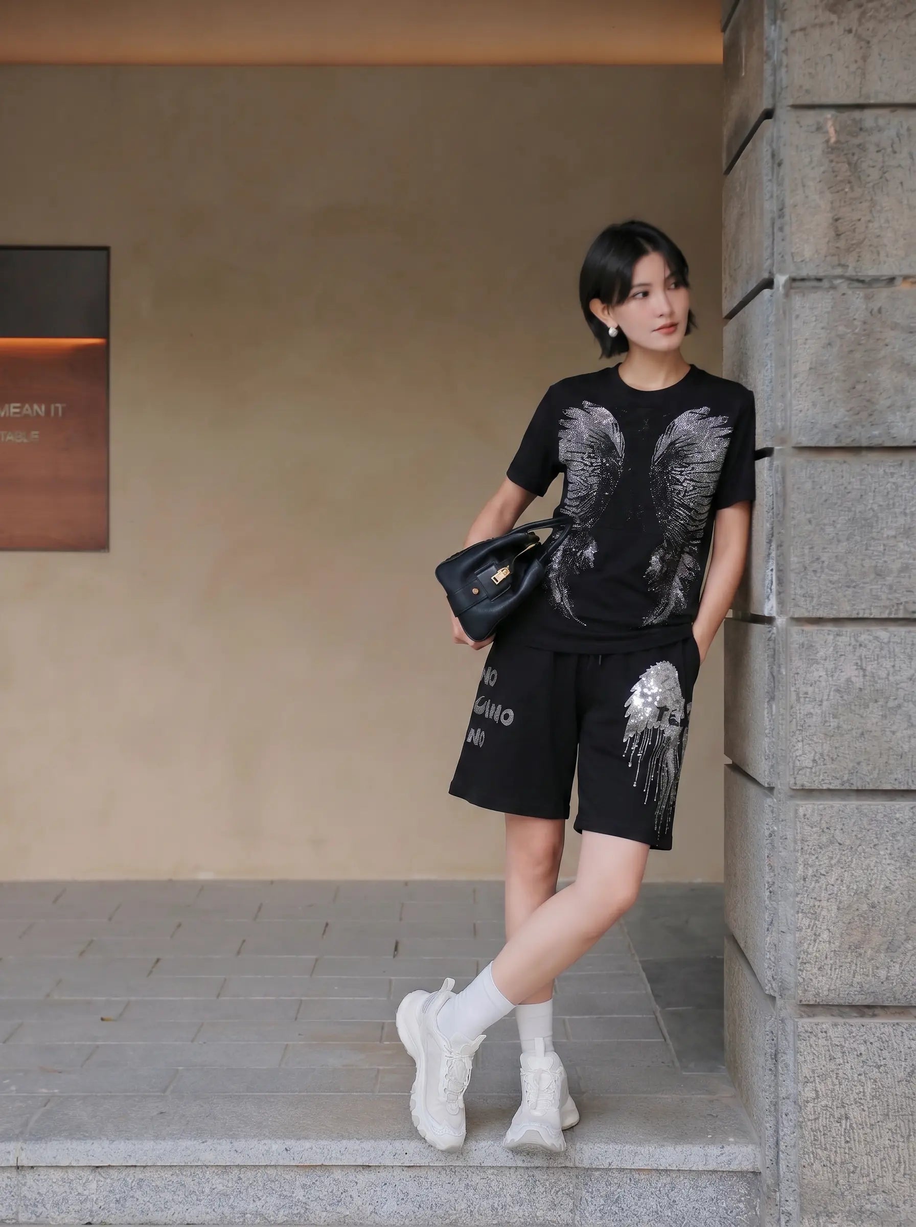 A woman stands outdoors against a stone pillar wearing a black graphic t-shirt and matching black shorts with white wing designs, white socks, and white sneakers, while holding a black handbag. TCH Rhinestone Angel Wing Graphic Shorts