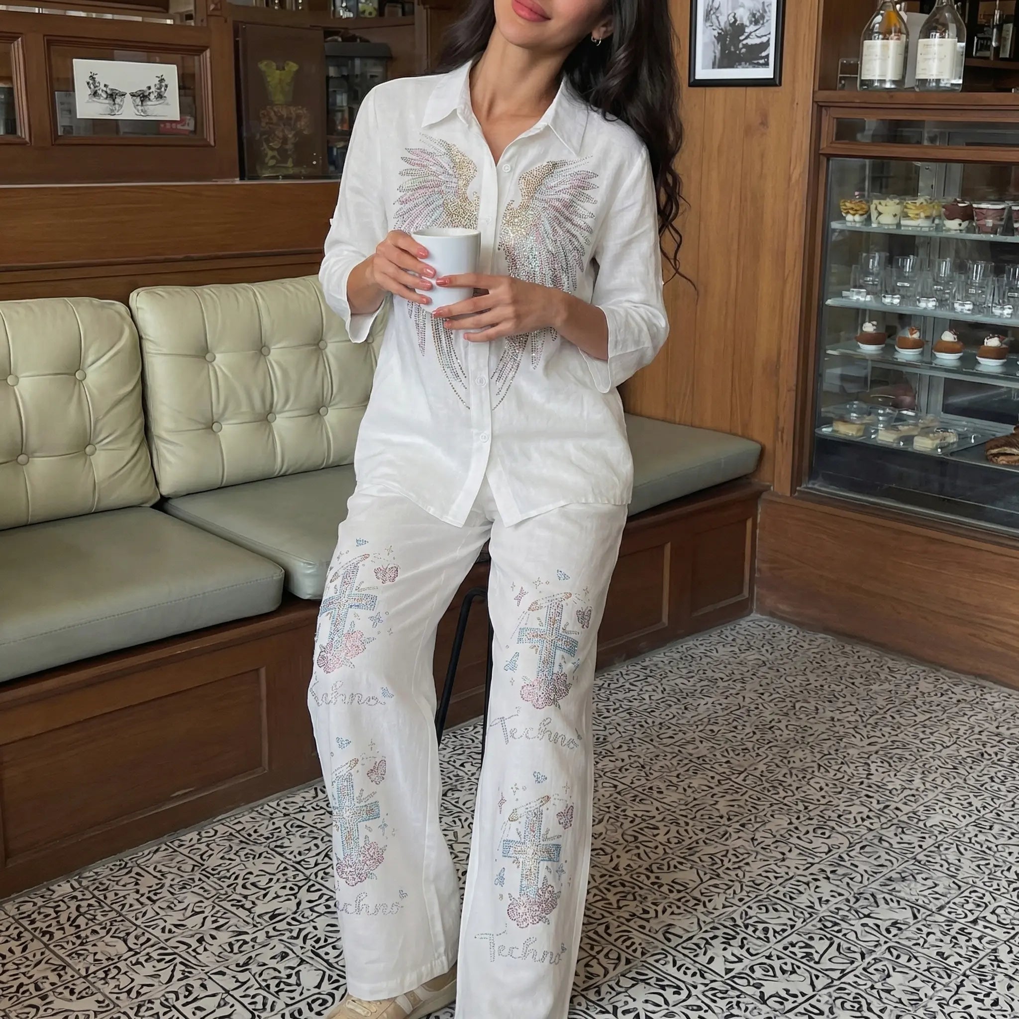 Woman in a white embroidered outfit holding a cup in a room with a couch and shelves. TCH Rhinestone Angel Wings & Cross Set