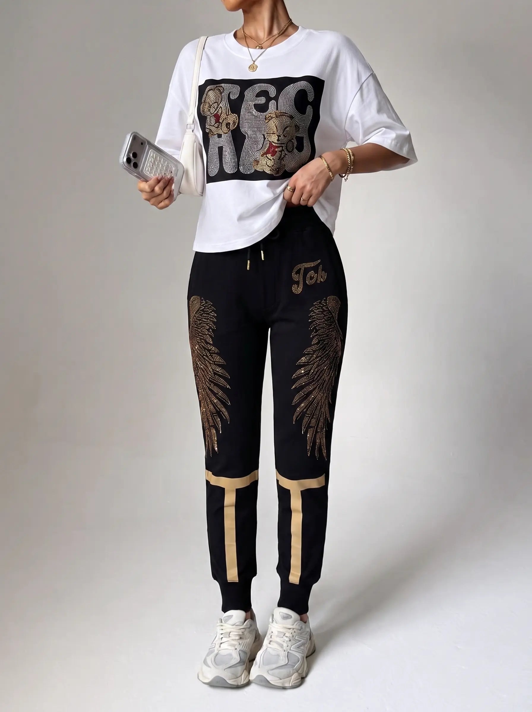 A model wearing a white graphic t-shirt and black jogger pants with gold wing designs and stripes, holding a phone against a neutral studio background. TCH Rhinestone Angel Wings Gold Detail Black Jogger Pants