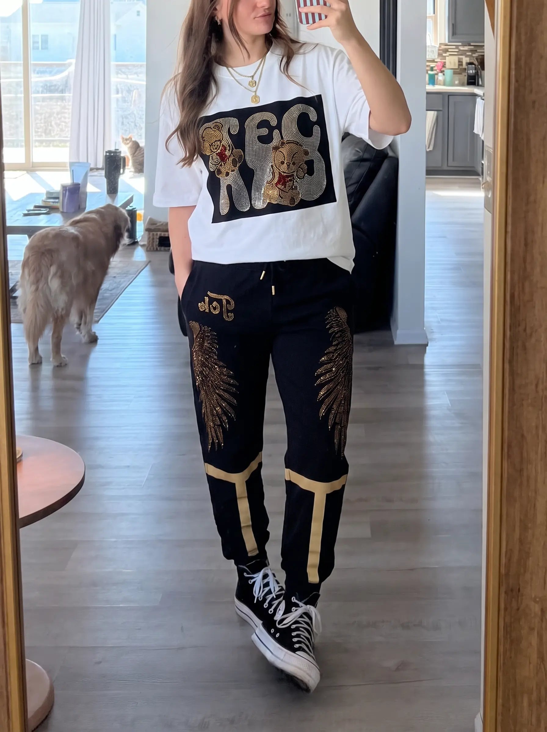 A woman takes a mirror selfie in a modern room, wearing a white graphic t-shirt with a black and gold design, black sweatpants with gold feather and stripe details, and black high-top sneakers. TCH Rhinestone Angel Wings Gold Detail Black Jogger Pants