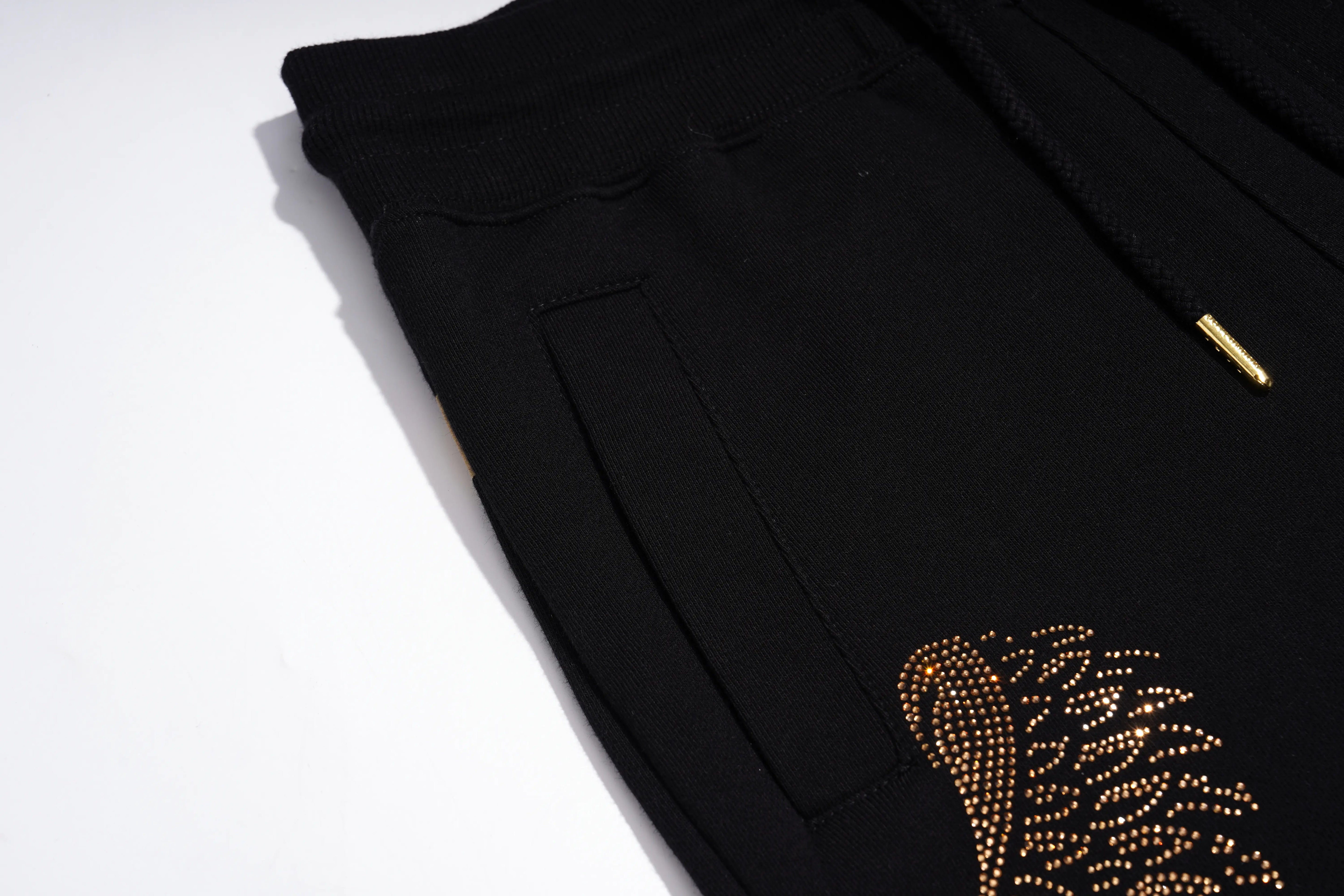 Close-up of black drawstring sweatpants with a gold rhinestone feather embellishment on the leg, shown on a white background. TCH Rhinestone Angel Wings Gold Detail Black Jogger Pants