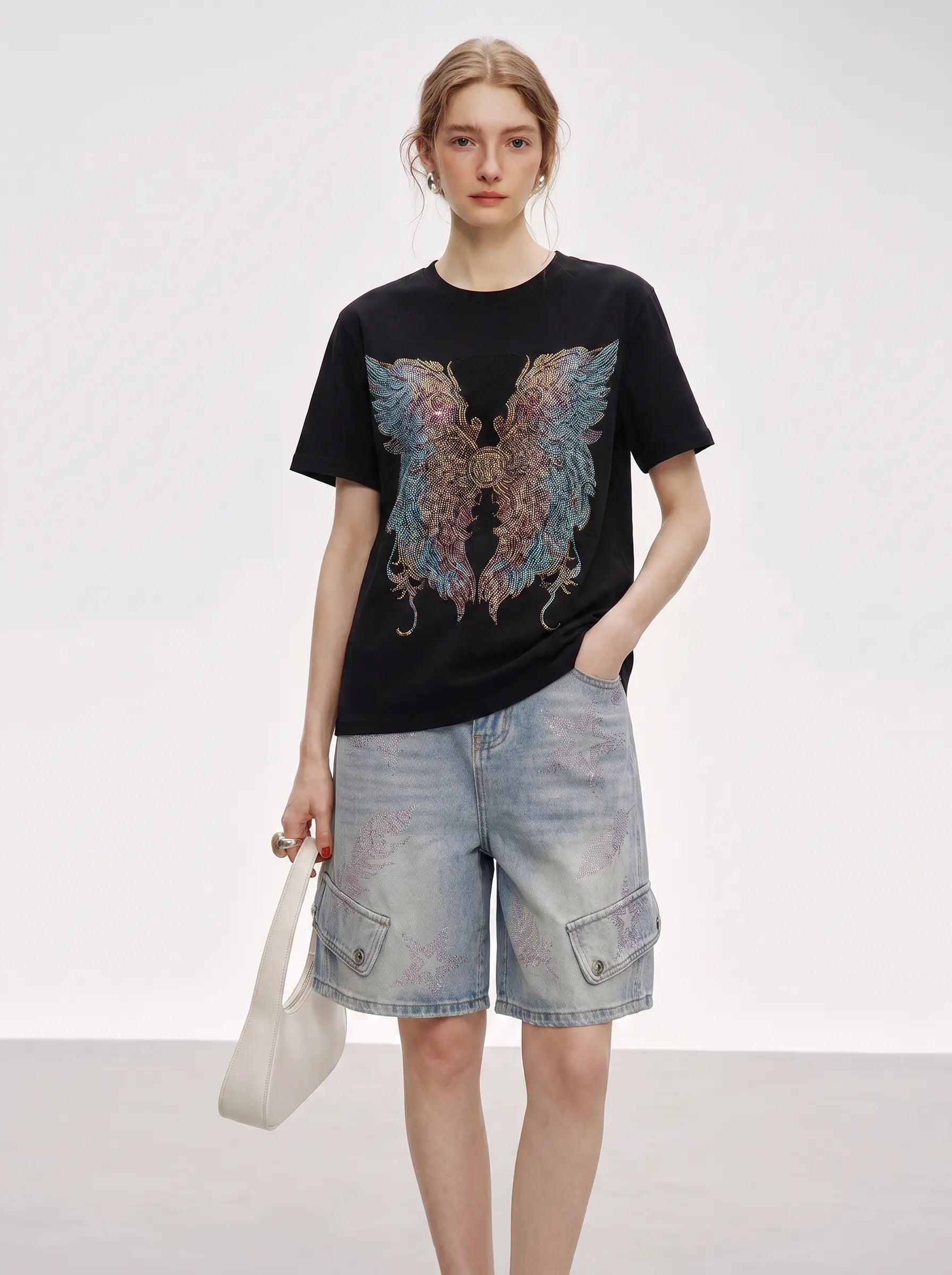 A model stands against a light gray studio background wearing a black short-sleeve T-shirt with a large multicolored wing graphic on the front, paired with light-wash denim cargo shorts and holding a white shoulder bag. TCH Rhinestone Angel Wings Graphic T-Shirt