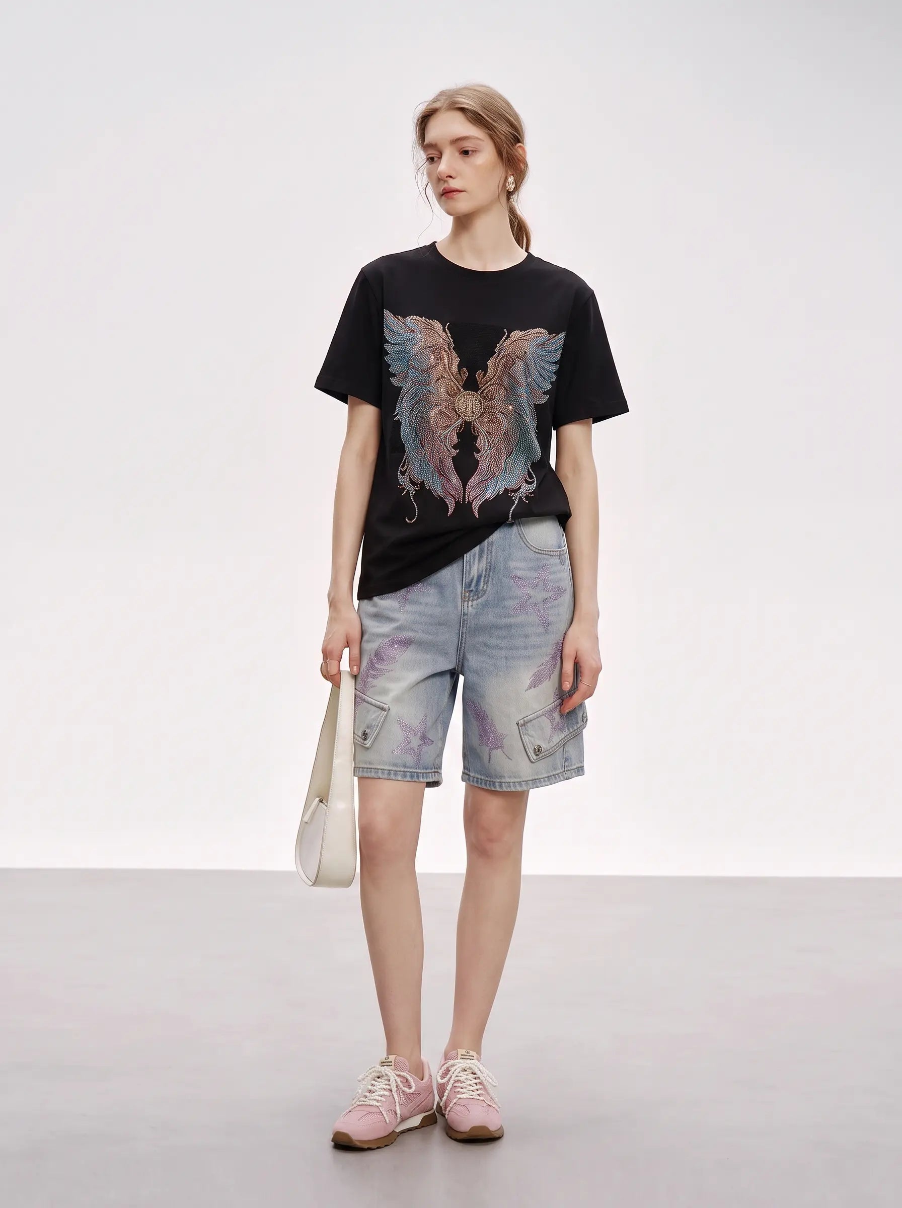 A model stands in a studio wearing a black graphic T-shirt with a large multicolor winged design, light-wash denim shorts, pink sneakers, and holding a cream shoulder bag. TCH Rhinestone Angel Wings Graphic T-Shirt