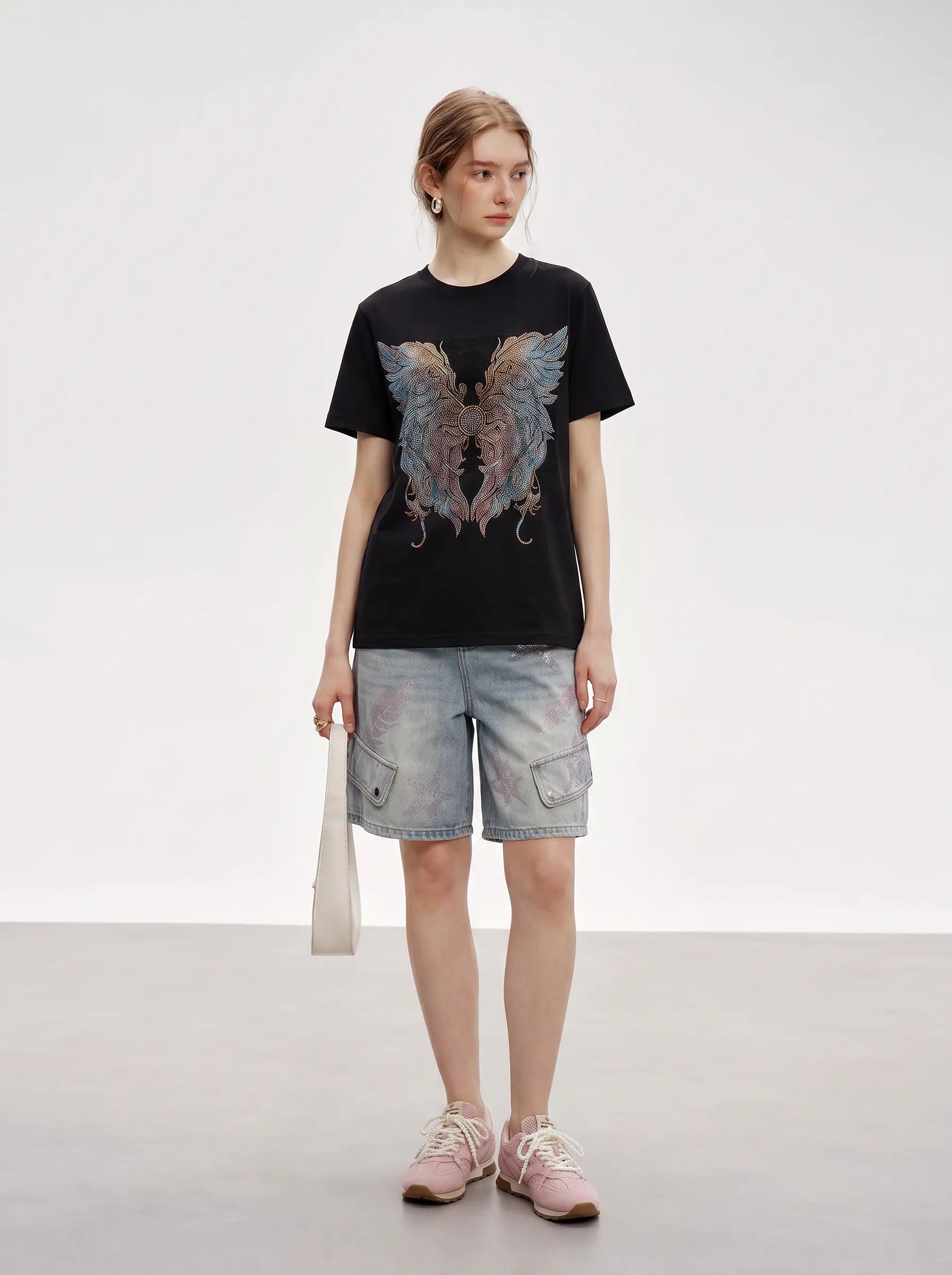 A model stands in a studio wearing a black graphic t-shirt with a large angel wing design, light-wash denim cargo shorts, and pink sneakers, holding a white shoulder bag. TCH Rhinestone Angel Wings Graphic T-Shirt