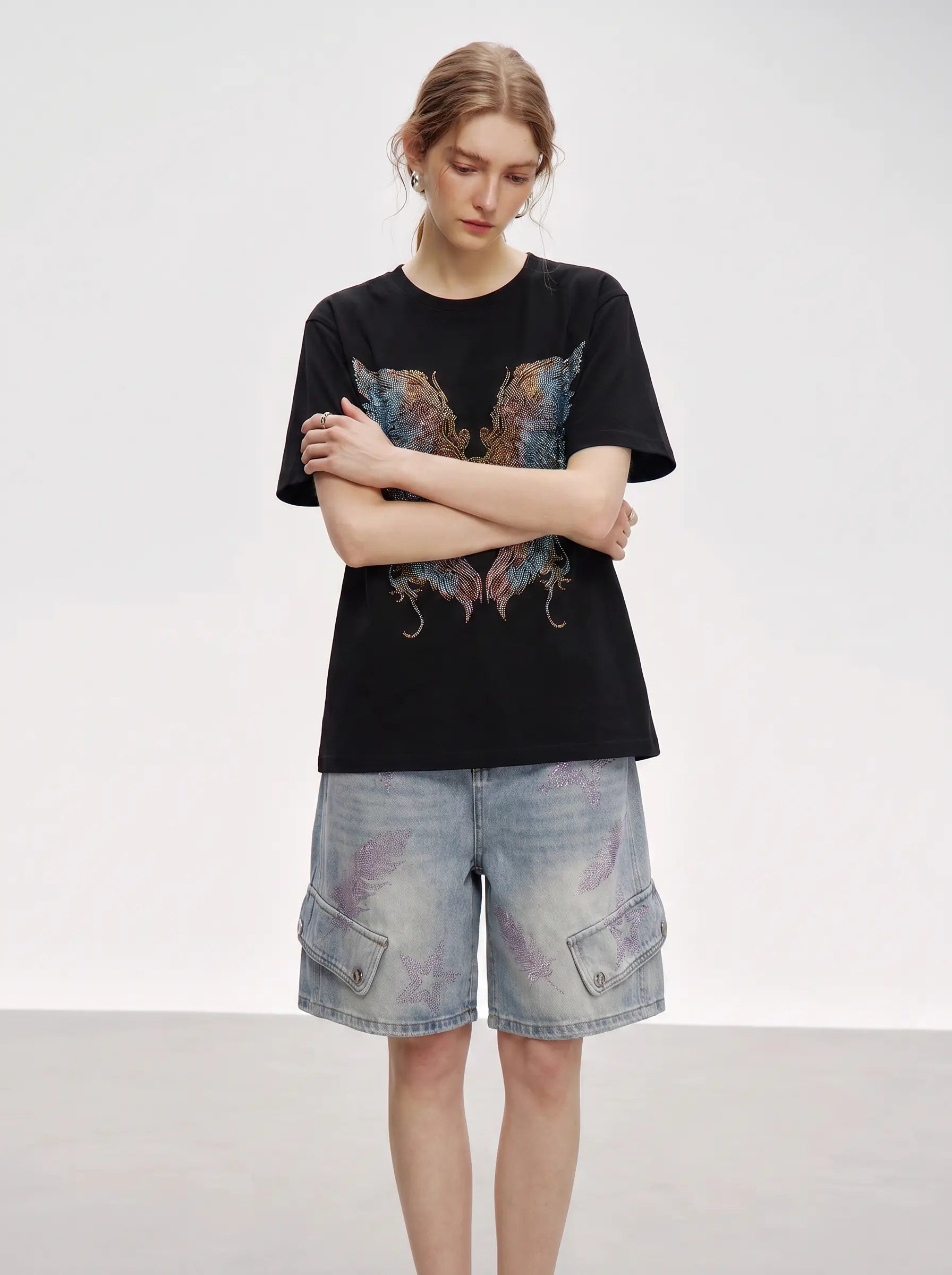 A woman modeling a black short-sleeve graphic T-shirt with a large multicolored butterfly print on the front, paired with light-wash denim cargo shorts, against a plain light gray studio background. TCH Rhinestone Angel Wings Graphic T-Shirt