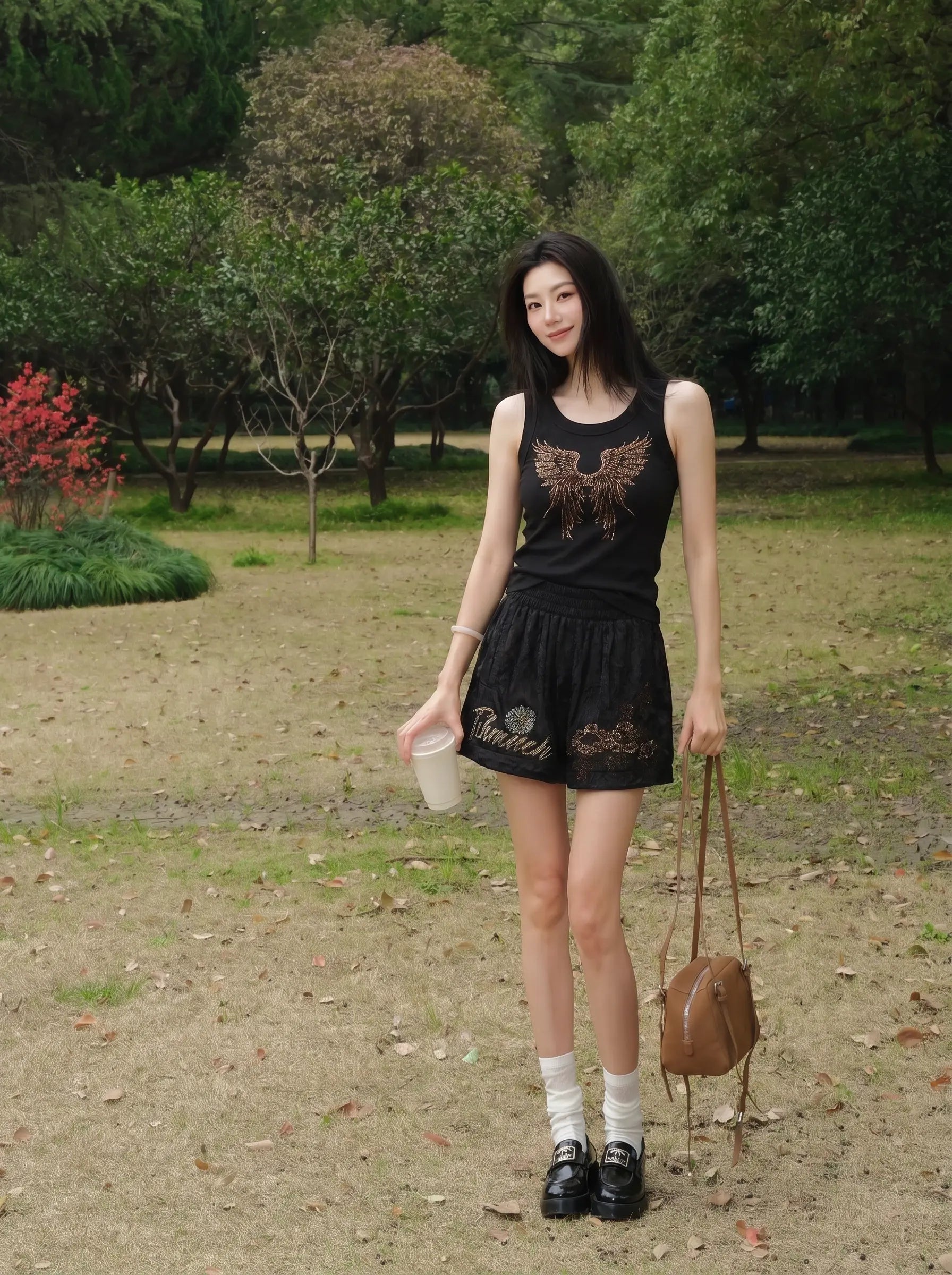 A woman stands outdoors in a park-like setting wearing a black sleeveless top and matching black shorts with brown graphic details. She is holding a light-colored cup in one hand and carrying a brown shoulder bag, with trees and grass in the background. TCH Rhinestone Angel Wings Ribbed Tank Top 