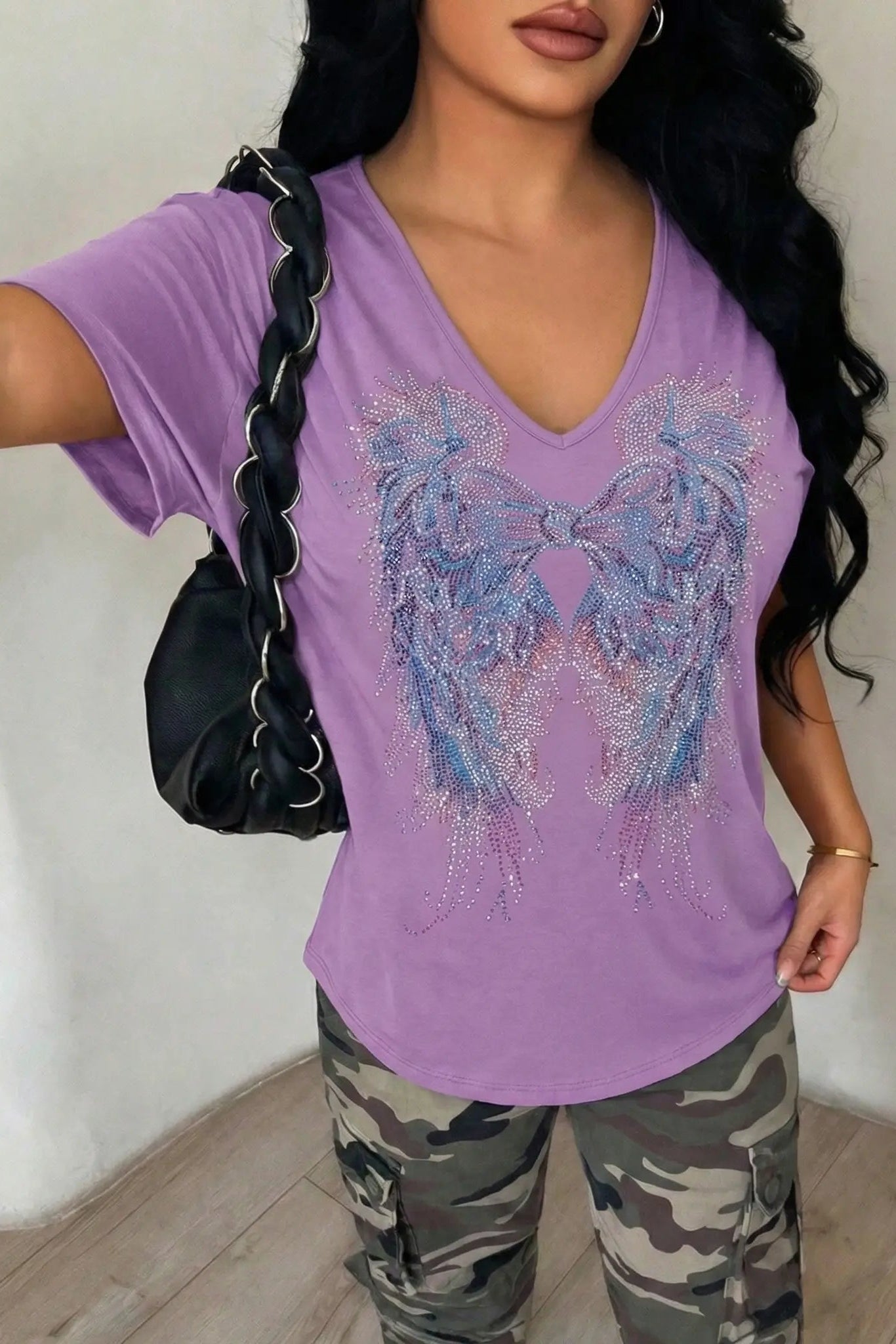 Woman wearing a purple t-shirt with a decorative bow design and camouflage pants, holding a black handbag. TCH Rhinestone Angel Wings V-Neck T-Shirt