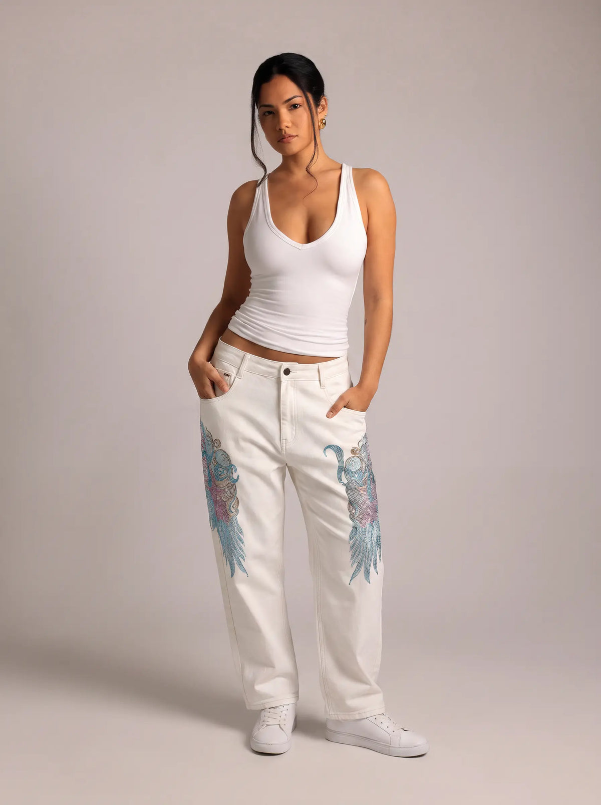A woman modeling white wide-leg jeans with colorful blue and pink wing graphics on the thighs, paired with a white ribbed tank top and white sneakers, against a solid light gray studio background. TCH Rhinestone Angel Wings White Straight-Leg Jeans