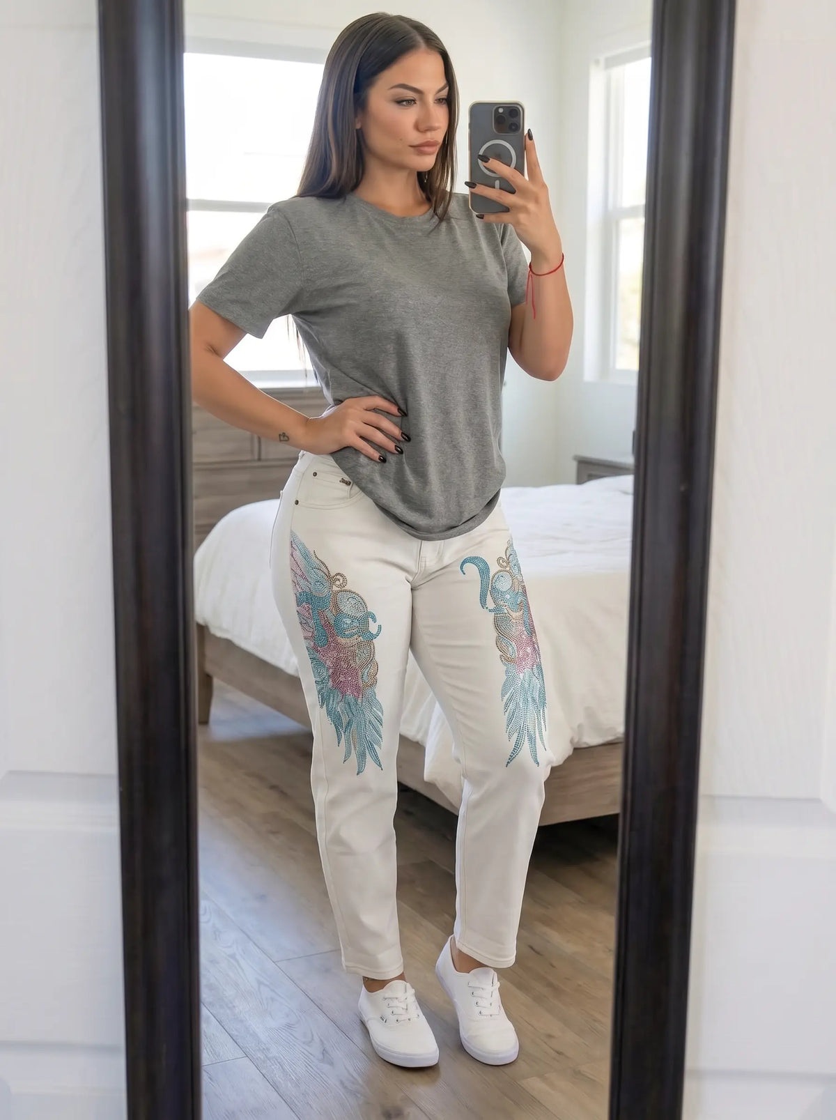 A woman takes a mirror selfie in a bedroom, wearing a heather gray short-sleeve t-shirt, white pants with colorful floral and feather graphics on the thighs, and white sneakers. TCH Rhinestone Angel Wings White Straight-Leg Jeans