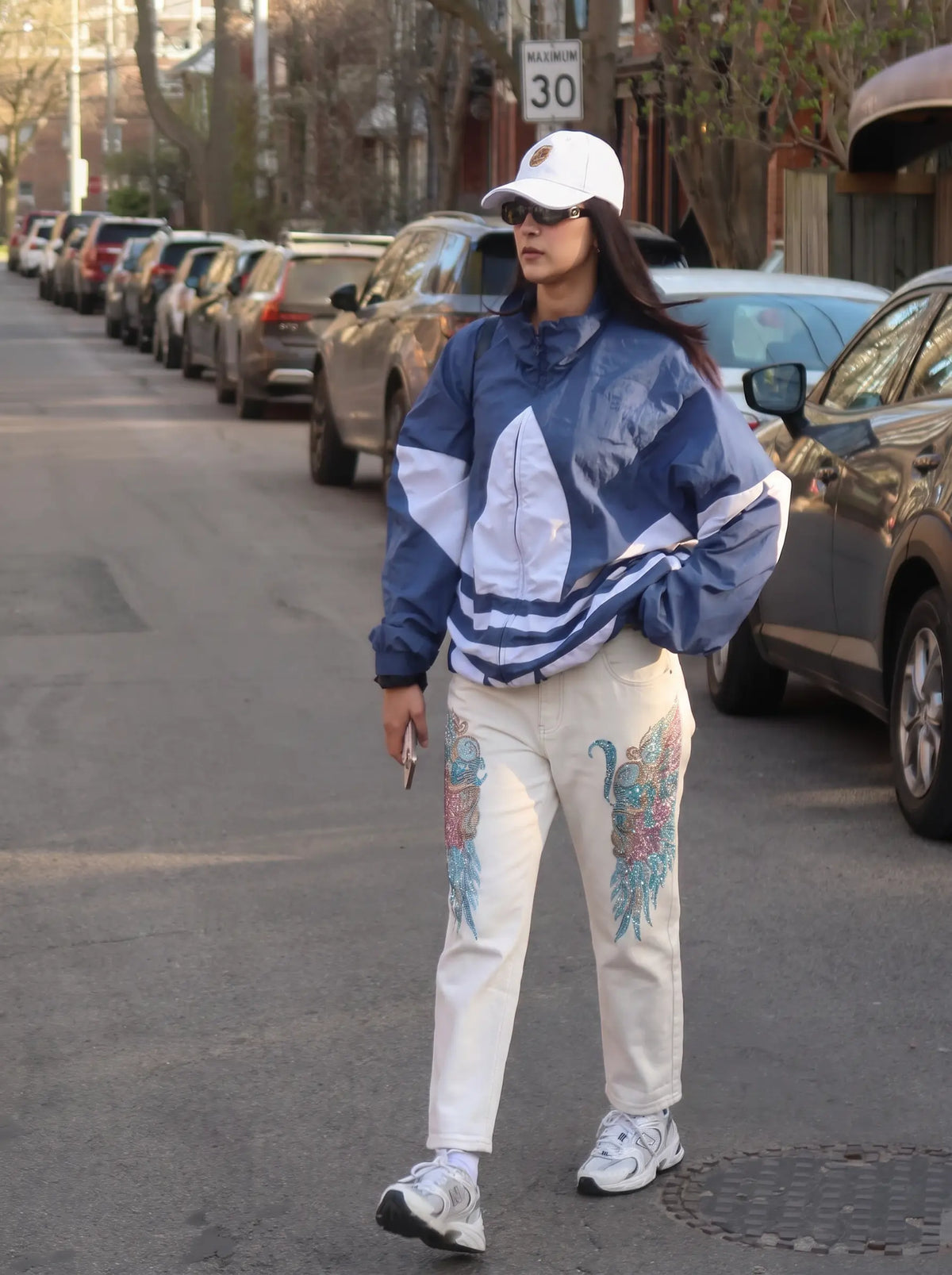 A woman stands on a city street wearing a blue and white windbreaker jacket, off-white embroidered pants, a white cap, and white sneakers. Cars are parked along the street in the background. TCH Rhinestone Angel Wings White Straight-Leg Jeans