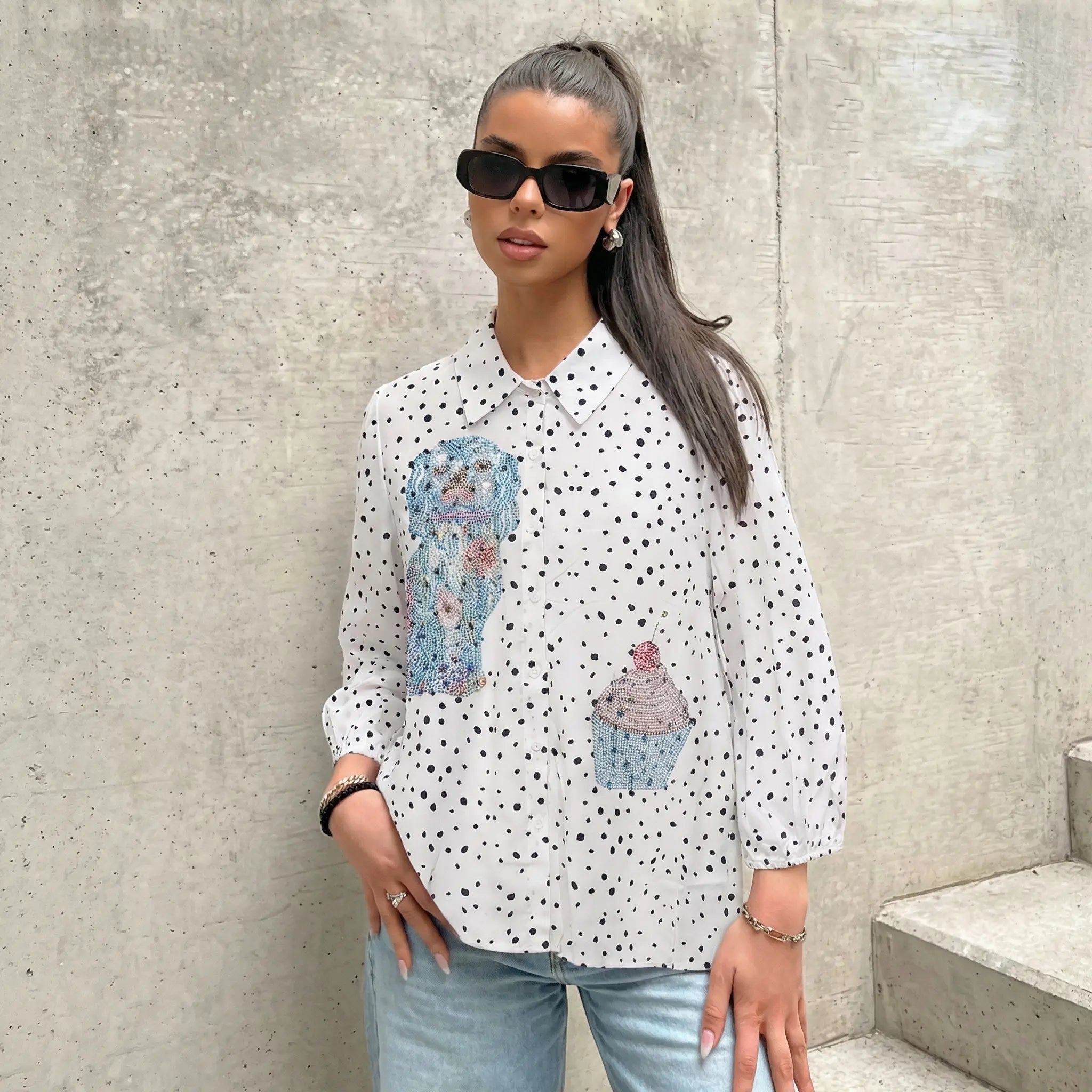 Woman wearing a white blouse with black polka dots and cupcake designs, standing against a concrete wall. TCH Rhinestone Animal Polka Dot Shirt