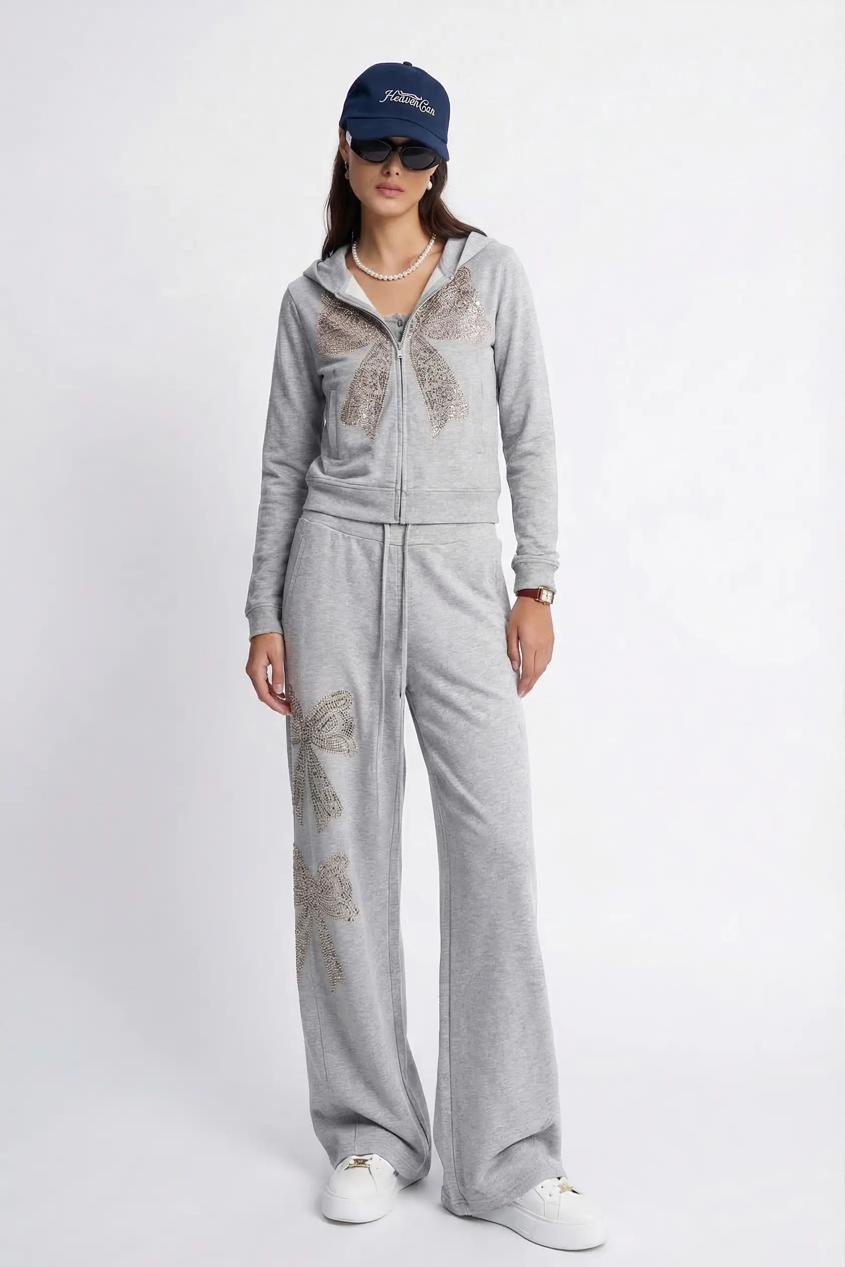 A model wearing a light gray zip-up hoodie and matching wide-leg sweatpants with metallic butterfly embellishments, styled with a navy baseball cap, sunglasses, a pearl necklace, and white sneakers against a plain white studio background. TCH Rhinestone Bow 3-Piece Lounge Set