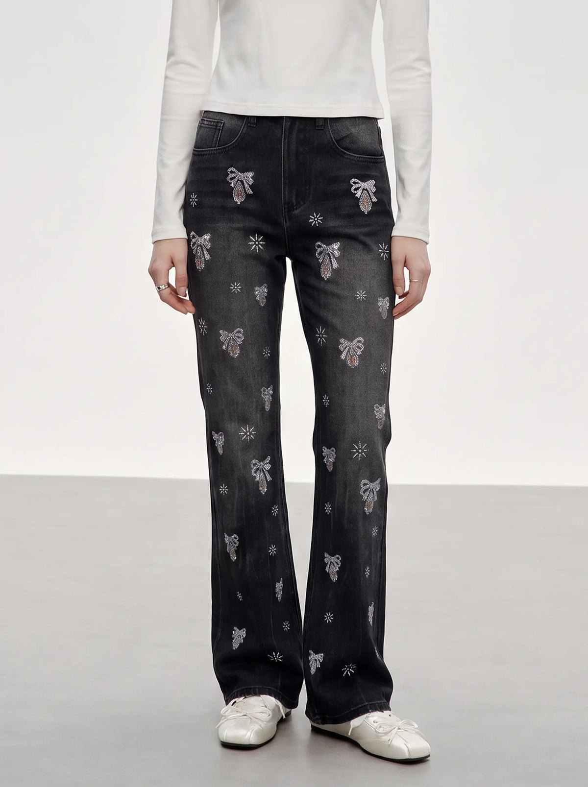 A model wearing a pair of dark gray wide-leg jeans with embroidered bows and floral motifs, paired with a white long-sleeve top and white sneakers. TCH Rhinestone Bow & Butterfly Flare Jeans