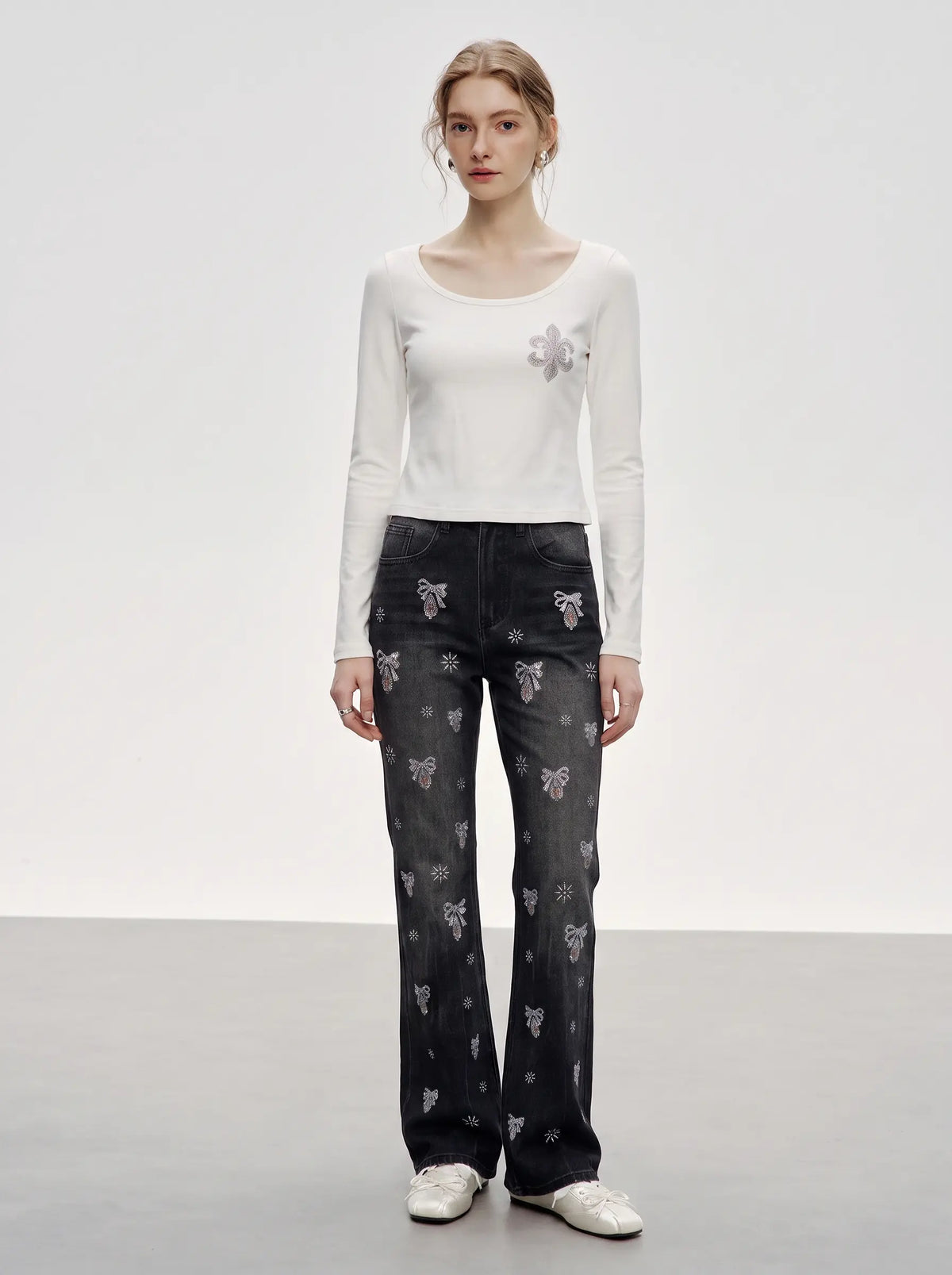 A model stands in a studio wearing a white long-sleeve top with a small floral patch on the chest, paired with dark gray wide-leg jeans featuring a repeating butterfly pattern, and white sneakers. TCH Rhinestone Bow & Butterfly Flare Jeans