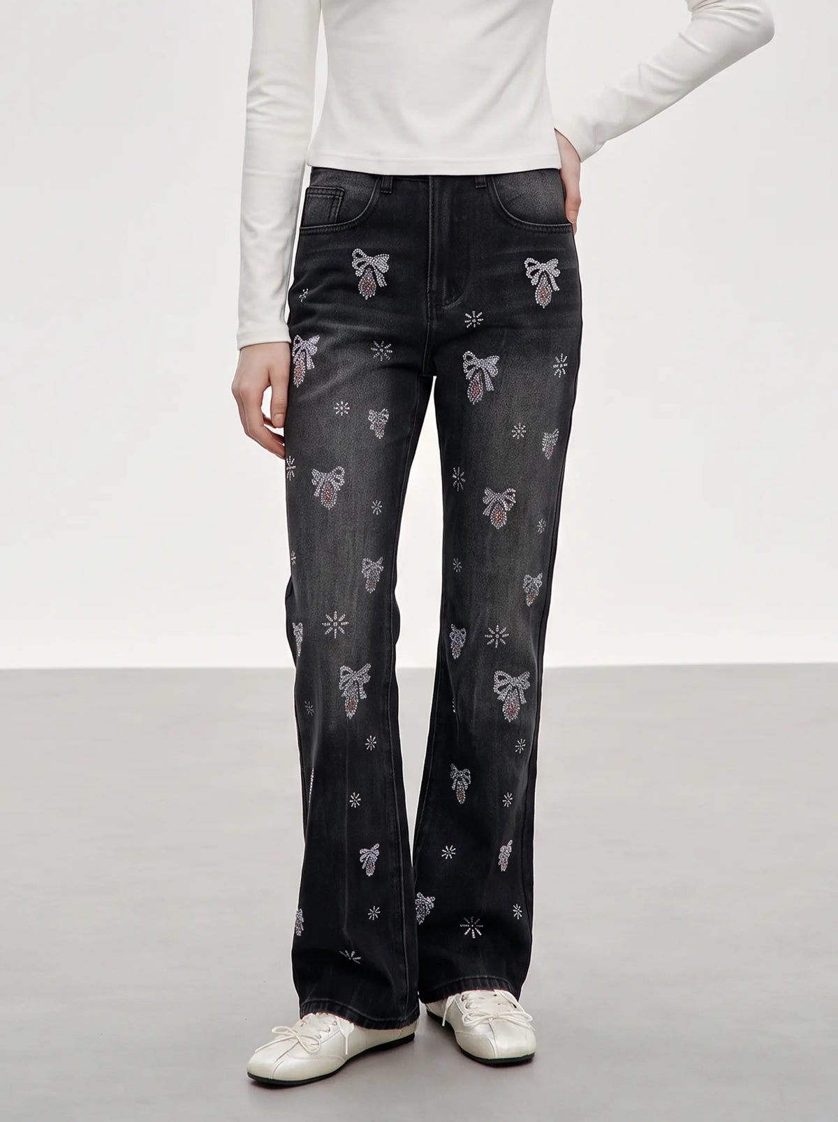 A model wearing dark gray denim flare jeans with embroidered bows and snowflakes, paired with a white long-sleeve top and white sneakers, shown from the waist down against a light gray studio background. TCH Rhinestone Bow & Butterfly Flare Jeans