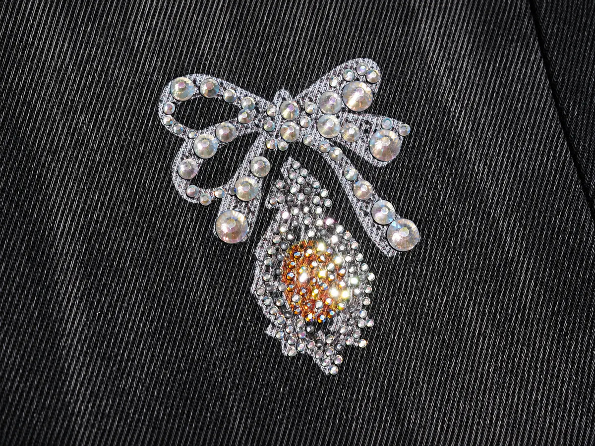 A silver-toned bow-shaped brooch embellished with clear rhinestones and pearl-like beads, featuring a teardrop-shaped lower section with a central cluster of warm amber-orange stones, displayed on a black textured fabric background. TCH Rhinestone Bow & Butterfly Flare Jeans