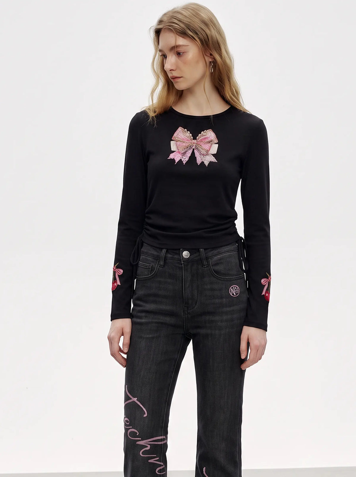 A model wearing a black long-sleeve crewneck top with a pink bow graphic on the chest and pink embroidery on the cuffs, paired with dark gray jeans. TCH Rhinestone Bow Cherry Drawstring Long Sleeve Top
