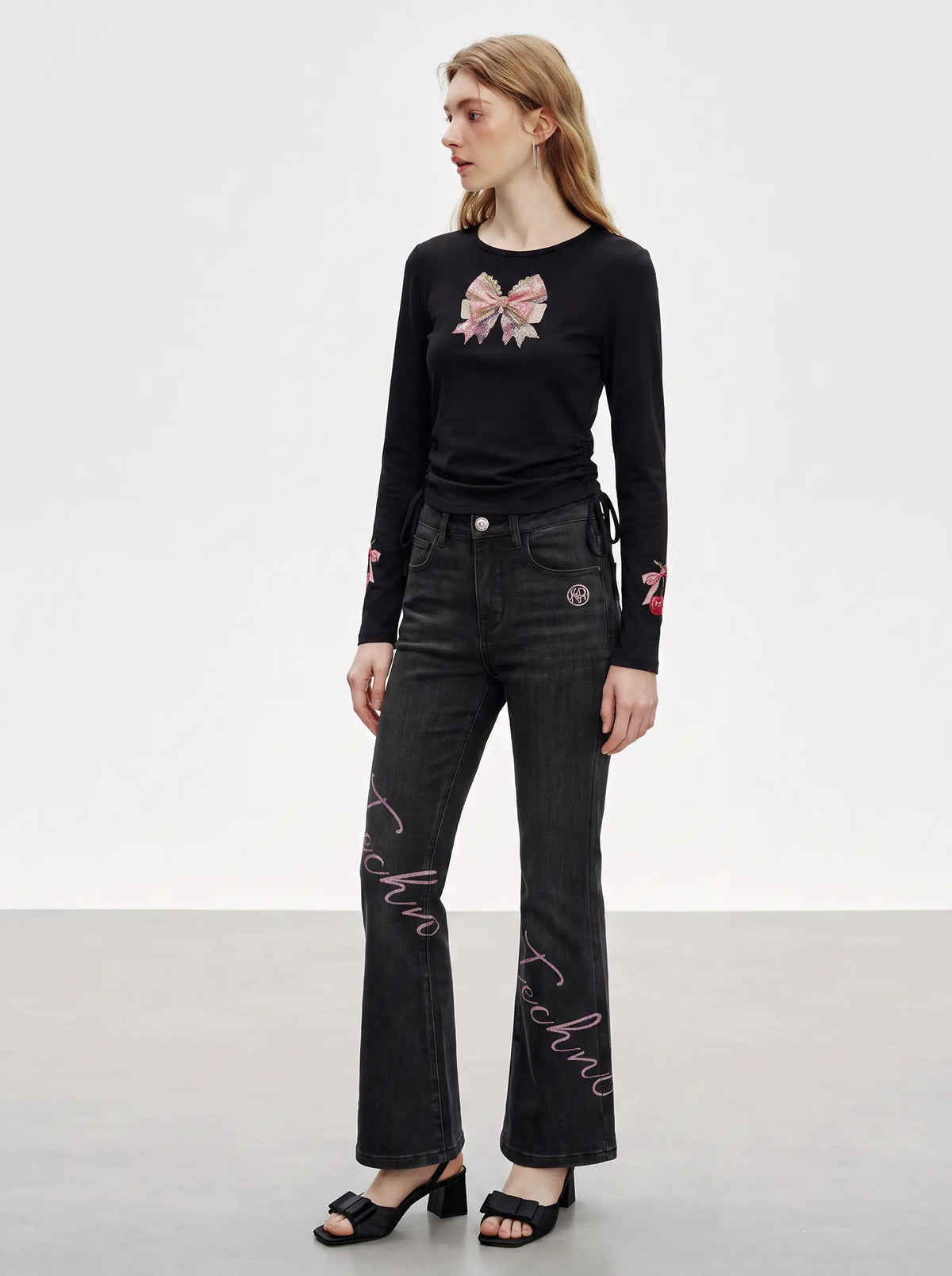A model wearing a black long-sleeve top with a pink bow graphic on the chest and dark gray flare jeans with pink script embroidery down the legs, paired with black heeled sandals. TCH Rhinestone Bow Cherry Drawstring Long Sleeve Top