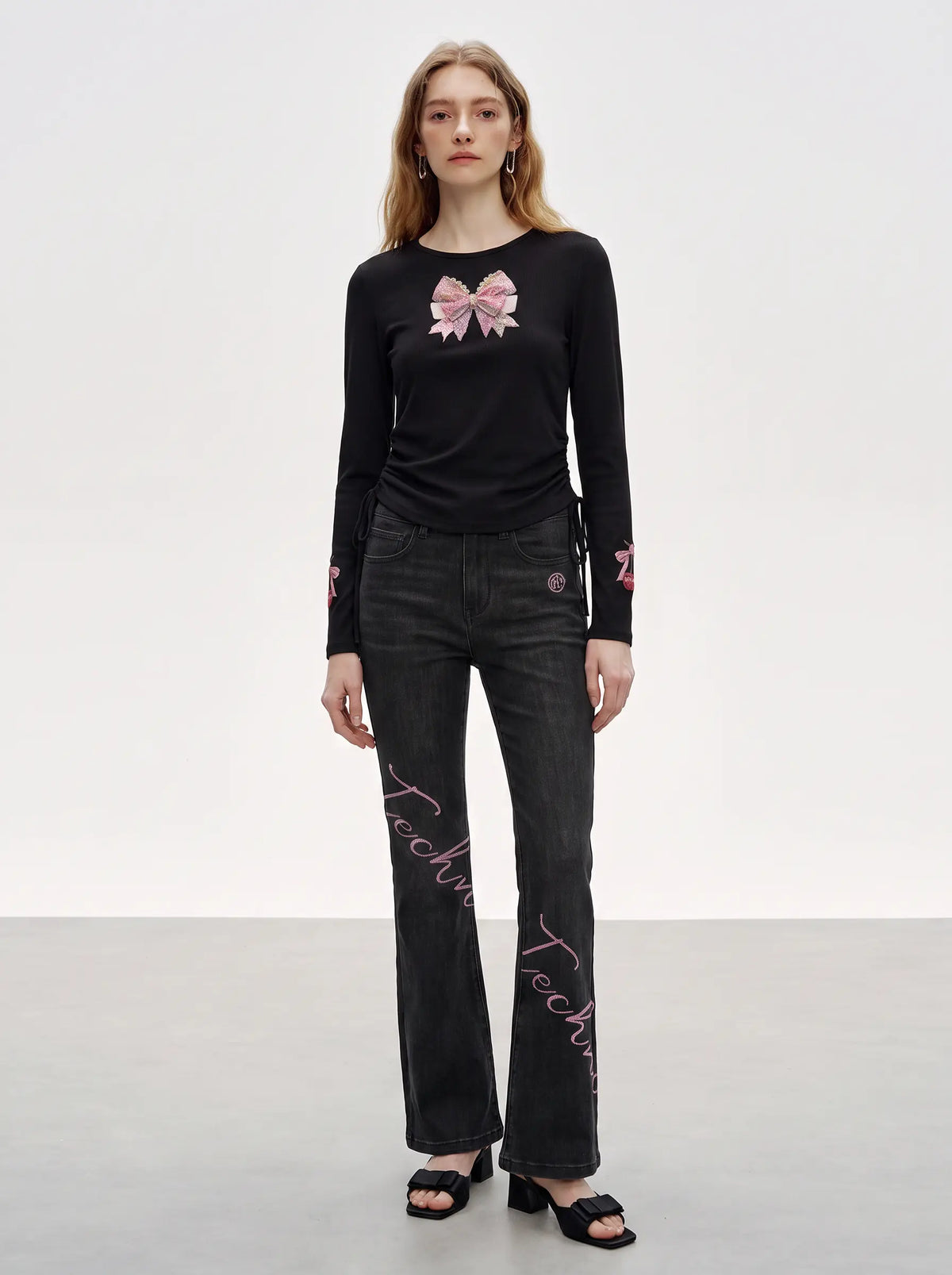 A model stands against a light gray studio background wearing a black long-sleeve top with a pink bow graphic on the chest and pink script embroidery on the jeans, paired with black heeled sandals. TCH Rhinestone Bow Cherry Drawstring Long Sleeve Top