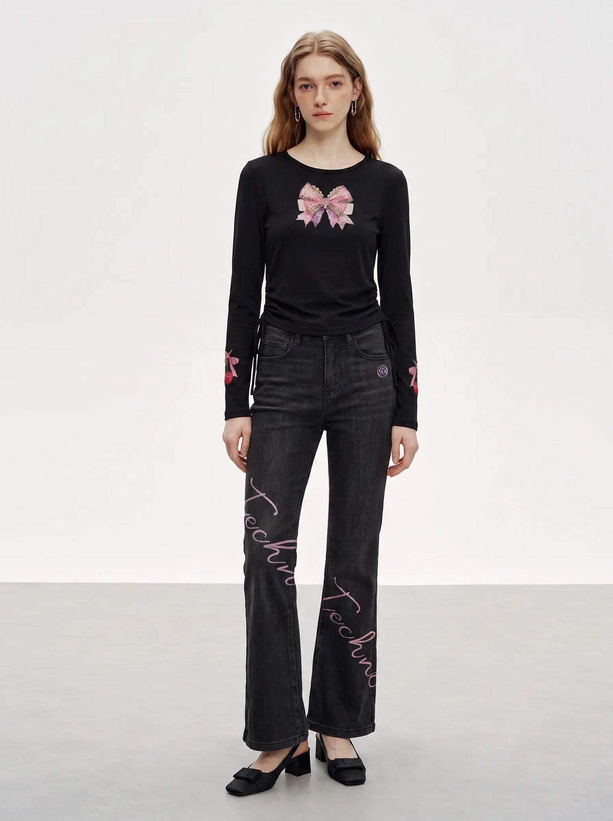A model stands in a studio wearing a black long-sleeve top with a pink bow graphic on the chest and pink script embroidery on the pants, paired with black jeans and black shoes. TCH Rhinestone Bow Cherry Drawstring Long Sleeve Top