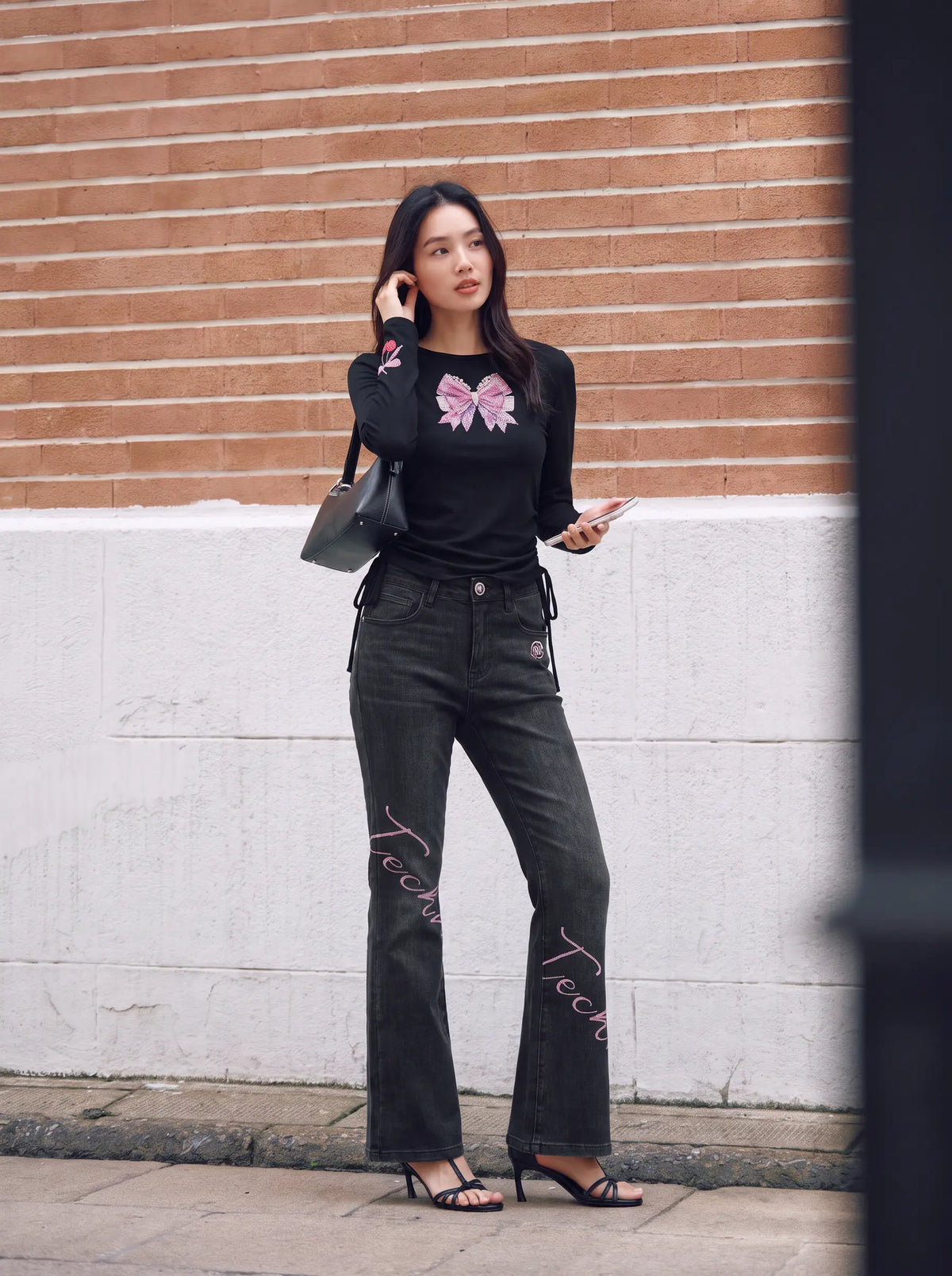 A woman stands outdoors against a brick and concrete wall wearing a black long-sleeve top with a pink bow graphic, black flared jeans with pink script embroidery, and black heeled sandals, while holding a black handbag. TCH Rhinestone Bow Cherry Drawstring Long Sleeve Top