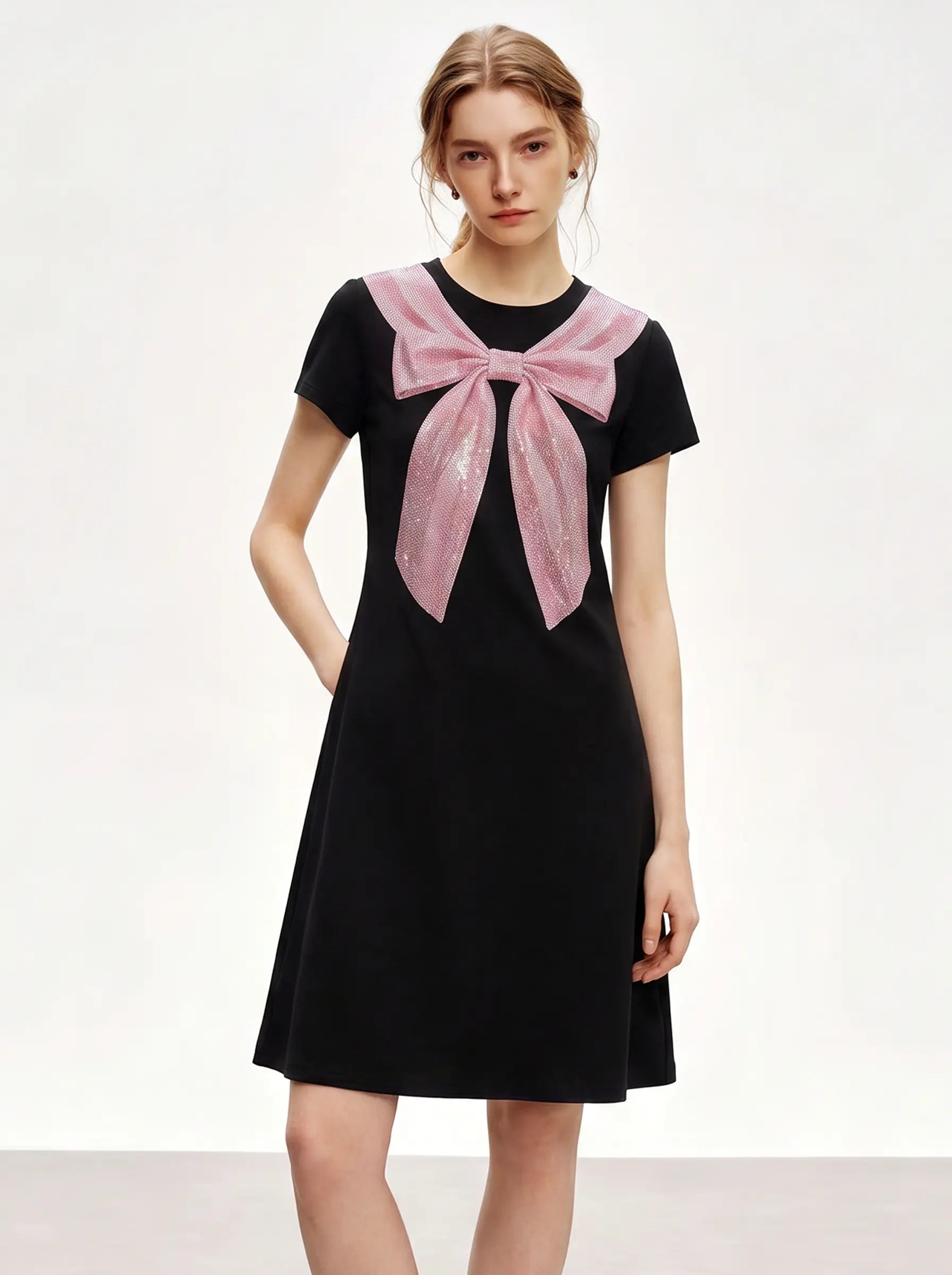 A woman modeling a black short-sleeve dress with a large pink bow detail across the chest, set against a plain light background. TCH Rhinestone Bow Collar Black Mini Dress