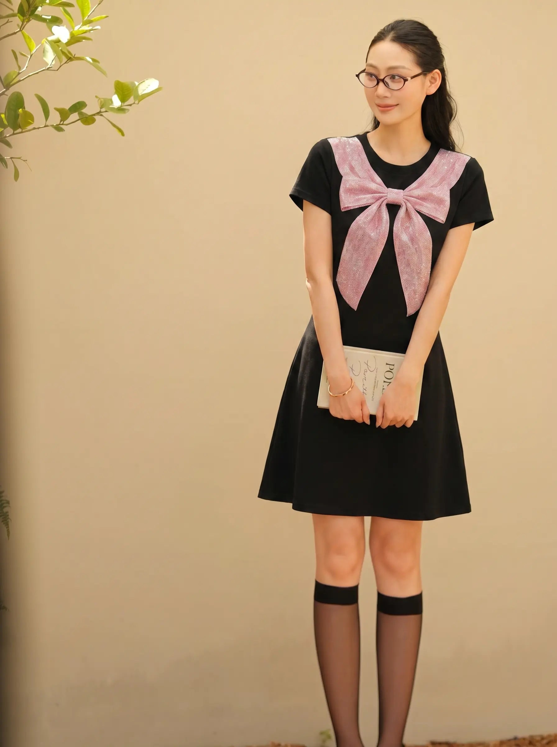 A woman wearing a black short-sleeve dress with a large pink bow detail on the chest, paired with black thigh-high stockings and holding a white clutch, standing against a beige wall. TCH Rhinestone Bow Collar Black Mini Dress