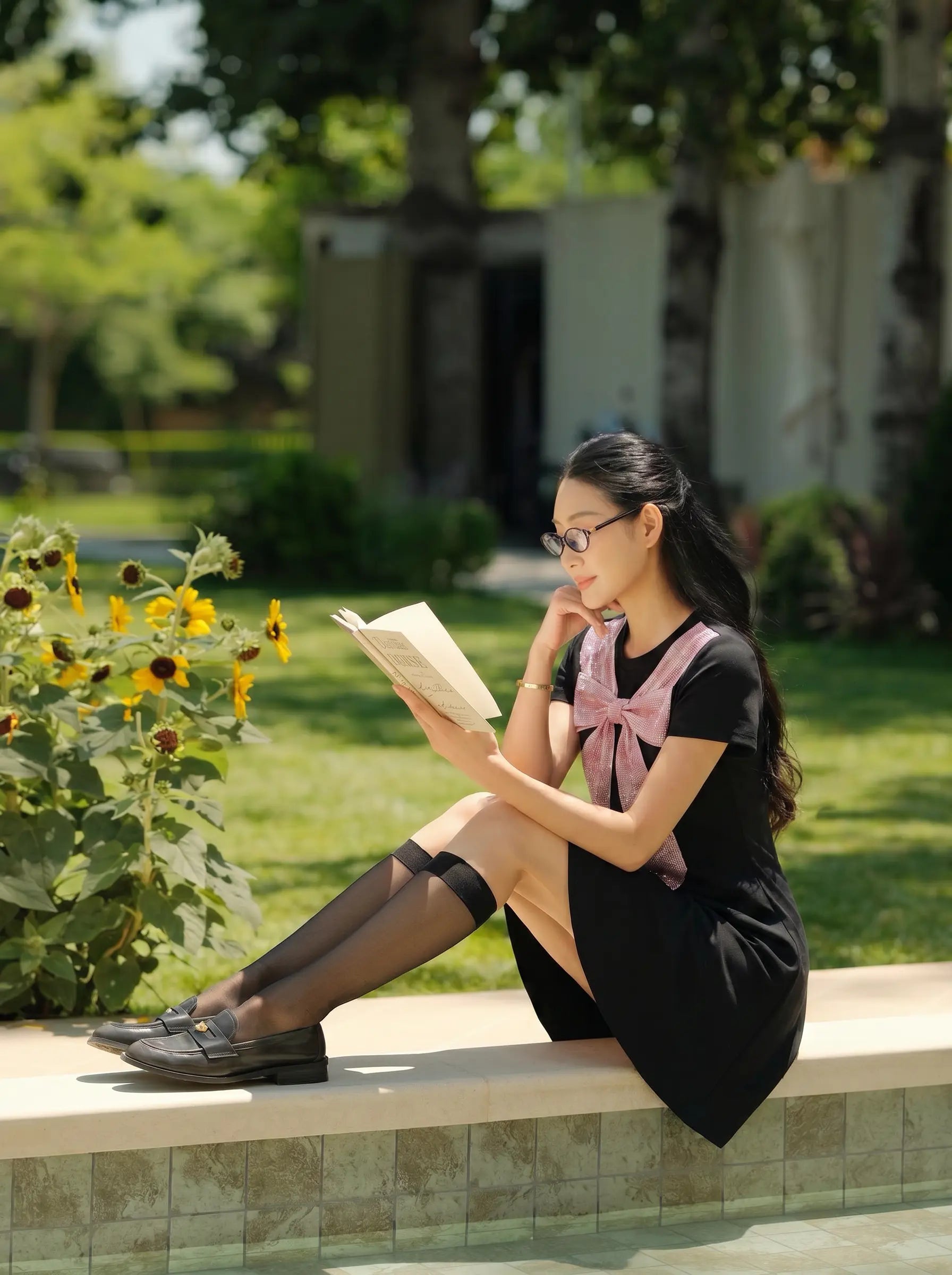 A woman wearing a black dress with a pink bow and glasses sits on a stone ledge in a sunny garden, reading a book. TCH Rhinestone Bow Collar Black Mini Dress