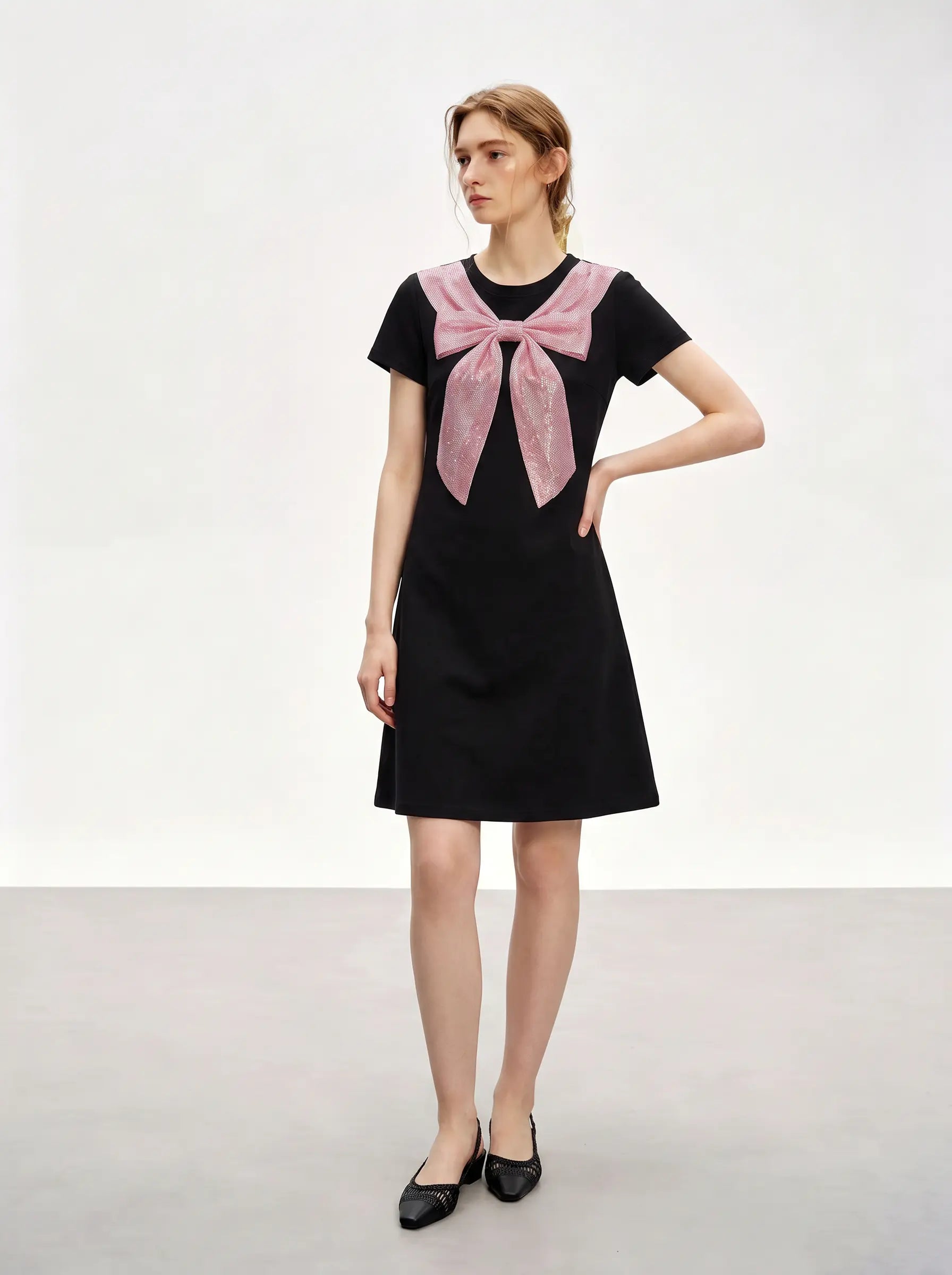 A woman modeling a black short-sleeve A-line dress with a large pink bow detail at the neckline, styled with black pointed flats against a plain light gray studio background. TCH Rhinestone Bow Collar Black Mini Dress