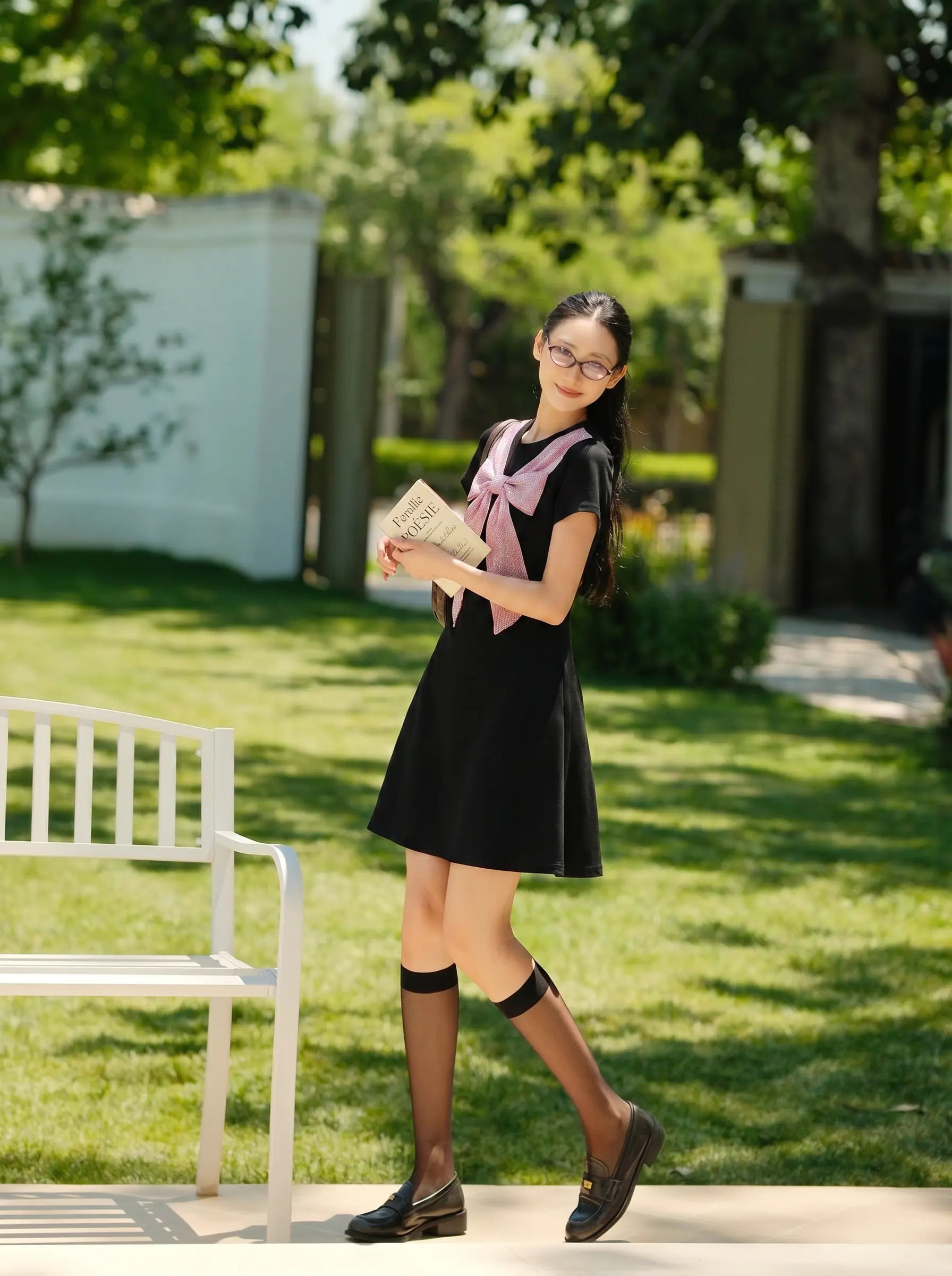 A young girl wearing a black short-sleeve dress with a pink bow tie, black knee-high socks, and dark loafers stands in a sunny garden holding a book. TCH Rhinestone Bow Collar Black Mini Dress