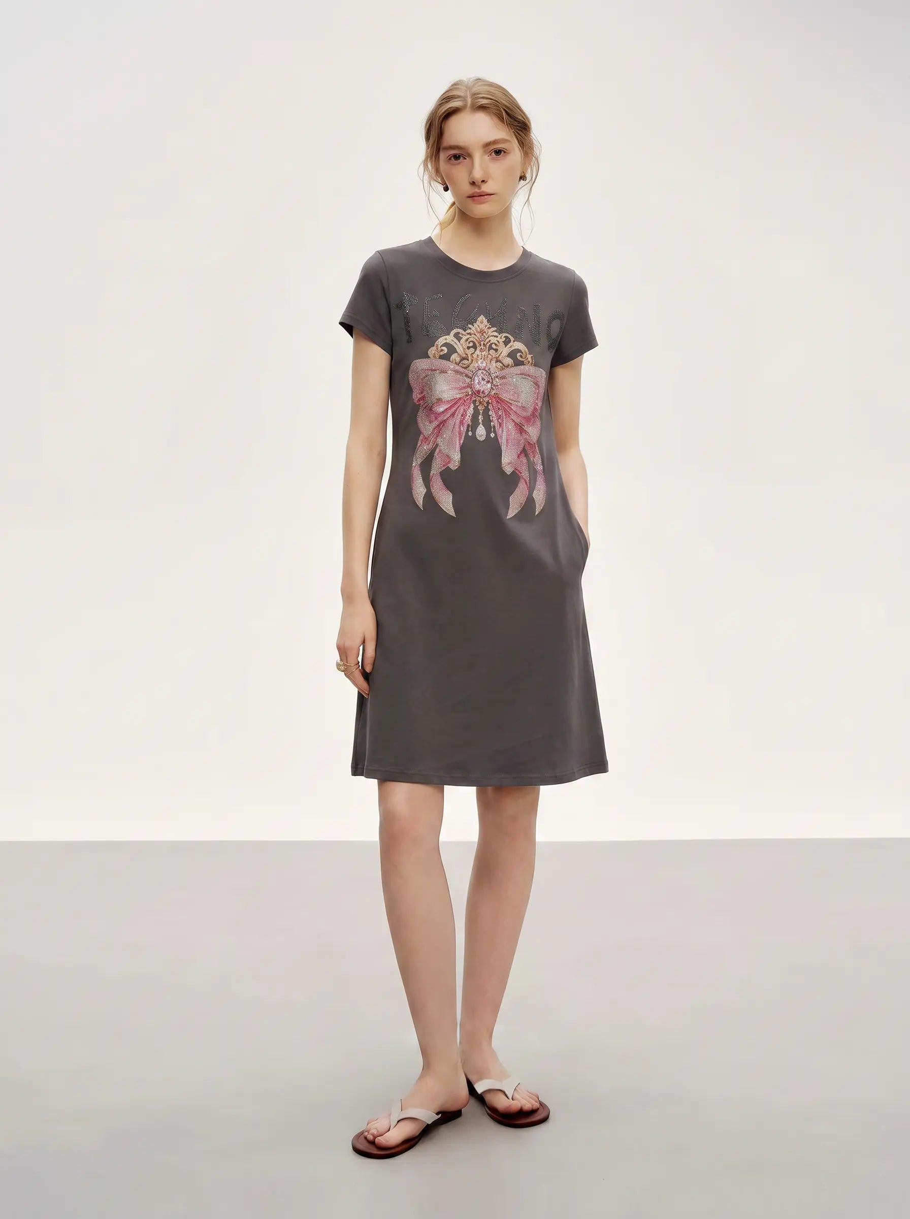 A woman modeling a dark gray short-sleeve t-shirt dress with a pink and gold graphic print on the front, paired with brown flip-flop sandals, set against a neutral studio background. TCH Rhinestone Bow Crown Graphic Mini Dress