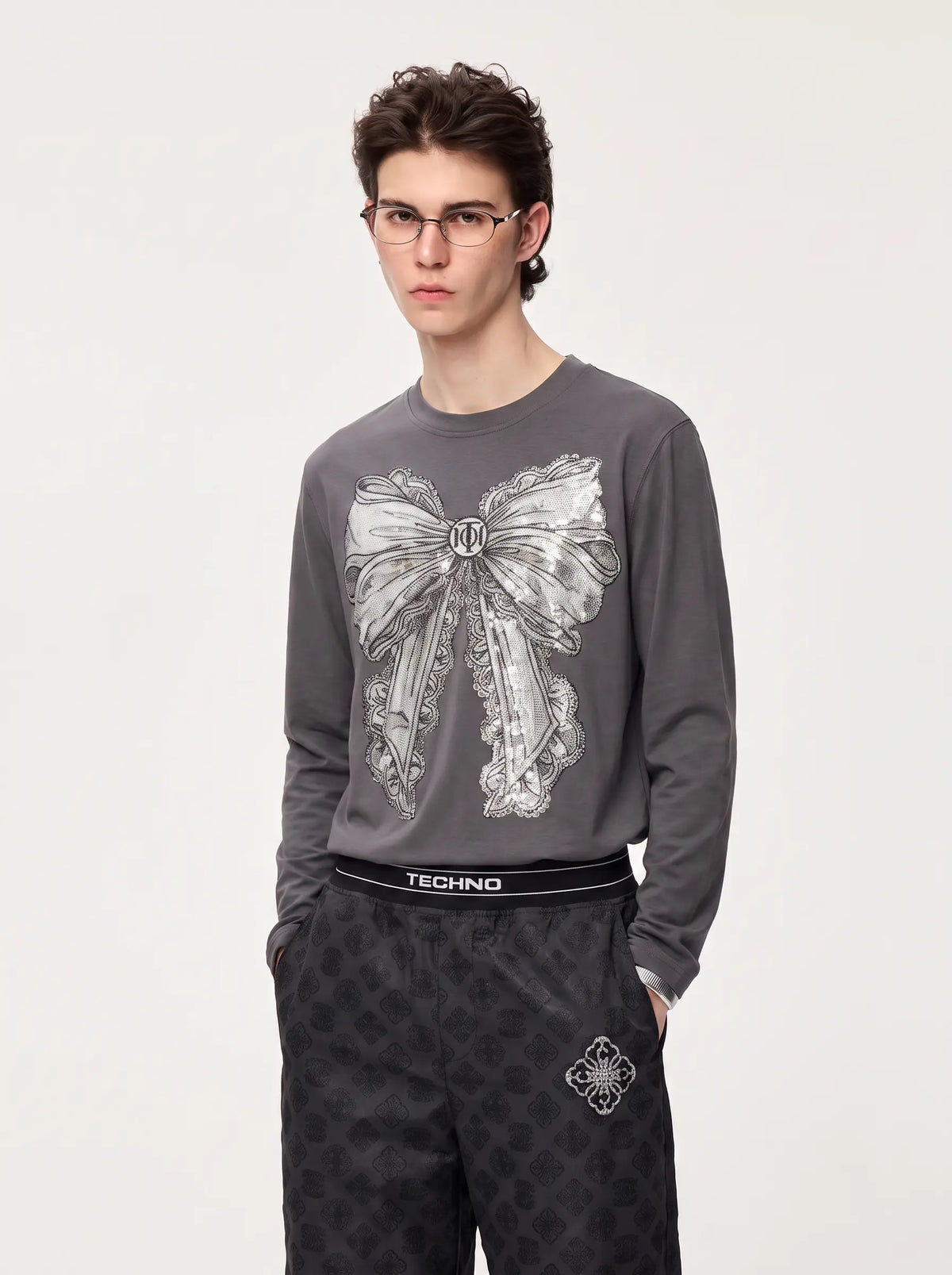 A young man wearing a dark gray long-sleeve sweatshirt with a large white bow graphic on the front, paired with dark patterned pants. The sweatshirt features a black waistband with the brand name 'TECHNO' printed on it. TCH Rhinestone Bow Graphic Fitted Long Sleeve Top