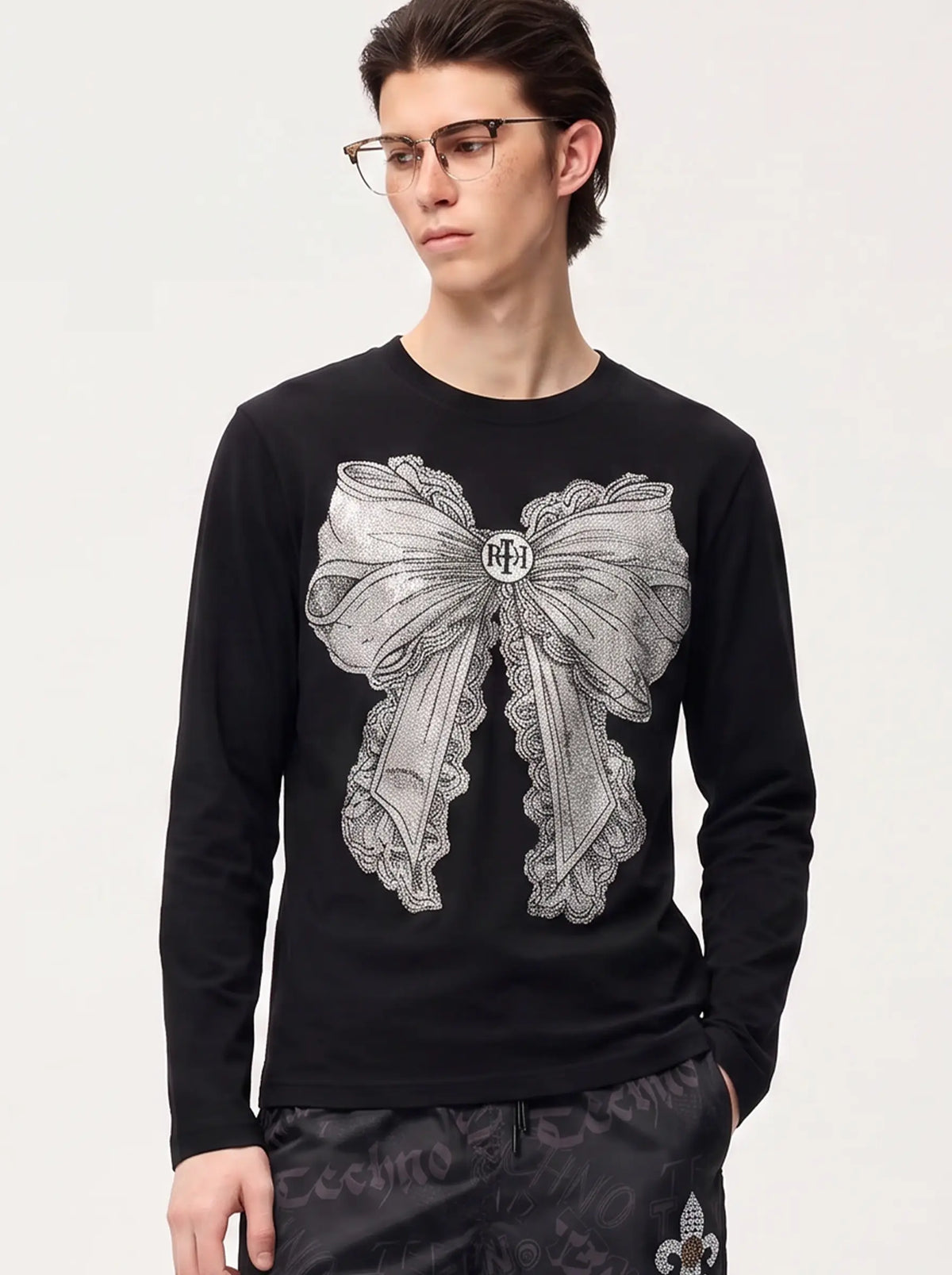 A model wearing a light blue denim jacket with various embroidered patches, including a teddy bear on the left chest and cherries on the sleeve, styled over a white t-shirt and dark pants. TCH Rhinestone Bow Graphic Fitted Long Sleeve Top