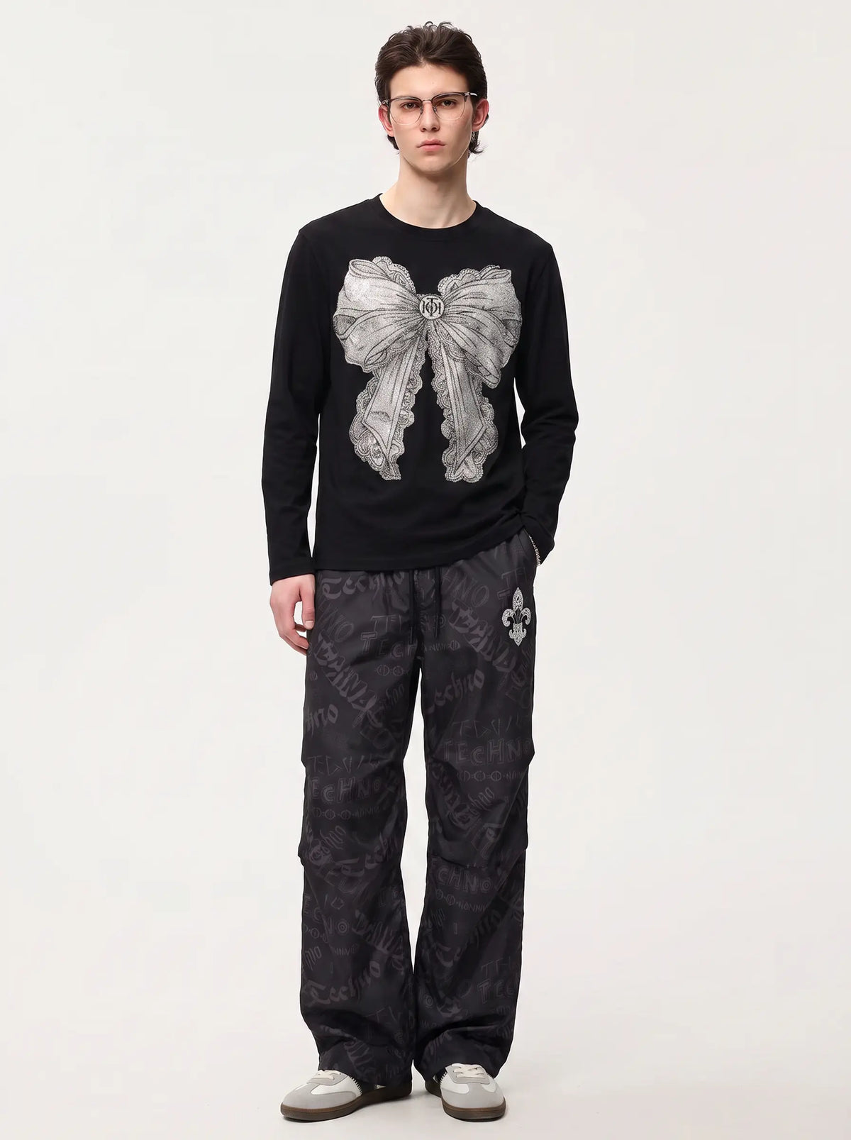 A model wearing a black long-sleeve crewneck shirt with a large gray bow graphic on the front, paired with dark patterned pants and gray shoes, standing against a plain light gray background. TCH Rhinestone Bow Graphic Fitted Long Sleeve Top