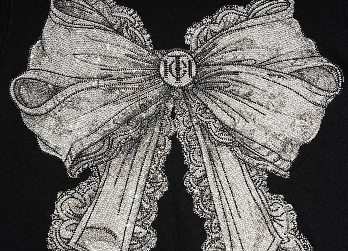 A close-up of a large decorative bow embellished with silver rhinestones and lace trim, centered on a black background. A circular monogram medallion is visible at the bow's center. TCH Rhinestone Bow Graphic Fitted Long Sleeve Top