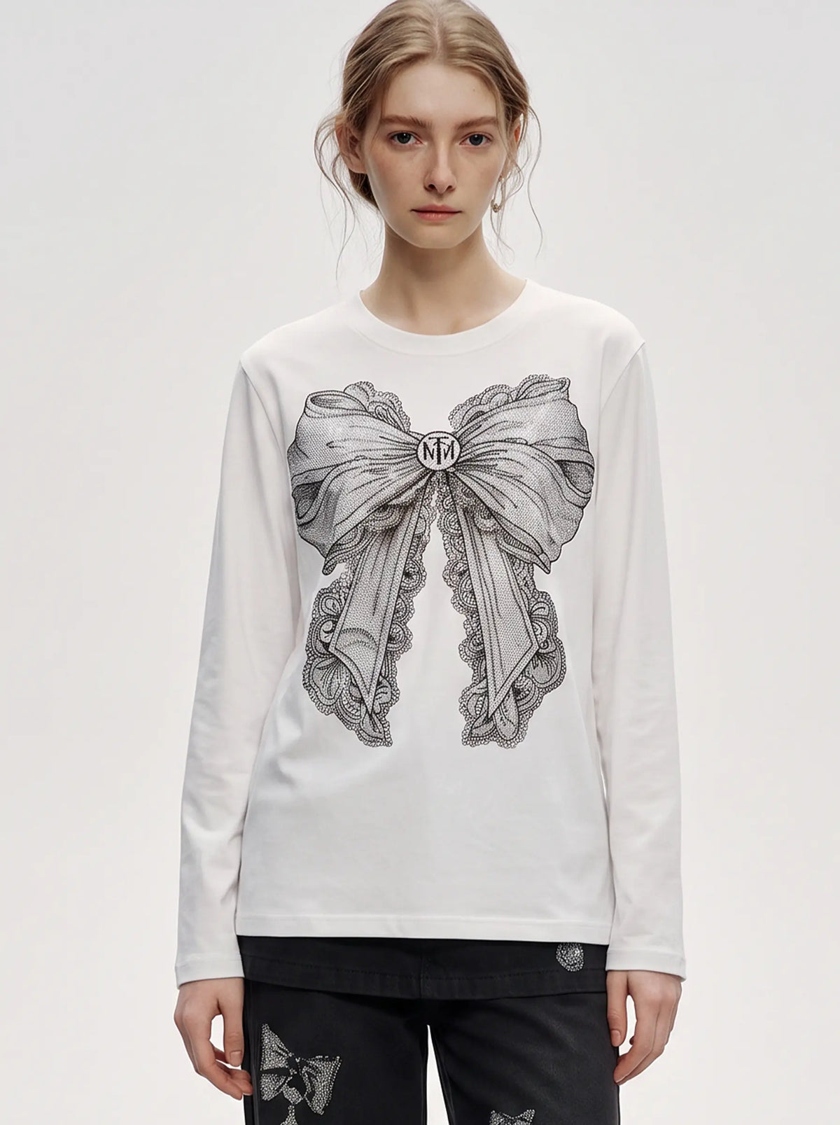 A model wearing a white long-sleeve t-shirt with a large black and gray bow graphic on the front, paired with black pants, against a plain light gray studio background. TCH Rhinestone Bow Graphic Fitted Long Sleeve Top
