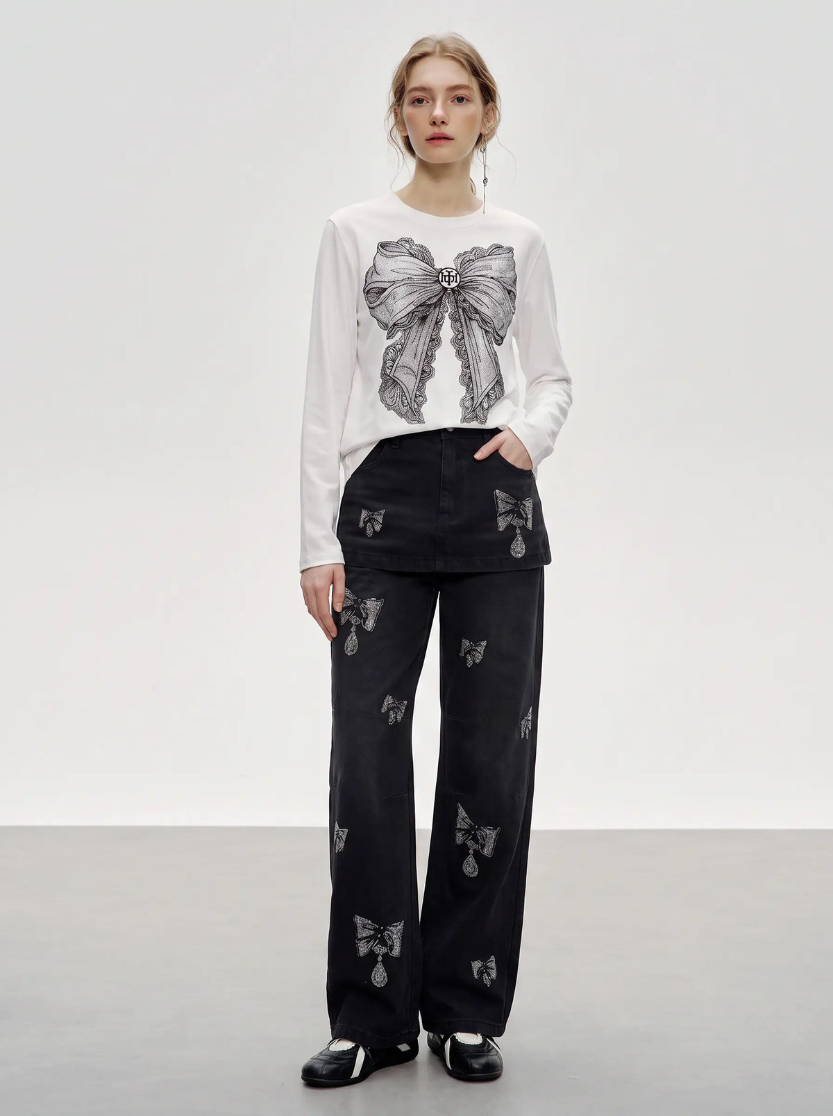 A model stands in a studio wearing a white long-sleeve top with a large gray bow graphic on the front, paired with black wide-leg pants featuring small gray bow motifs, and black shoes. TCH Rhinestone Bow Graphic Fitted Long Sleeve Top