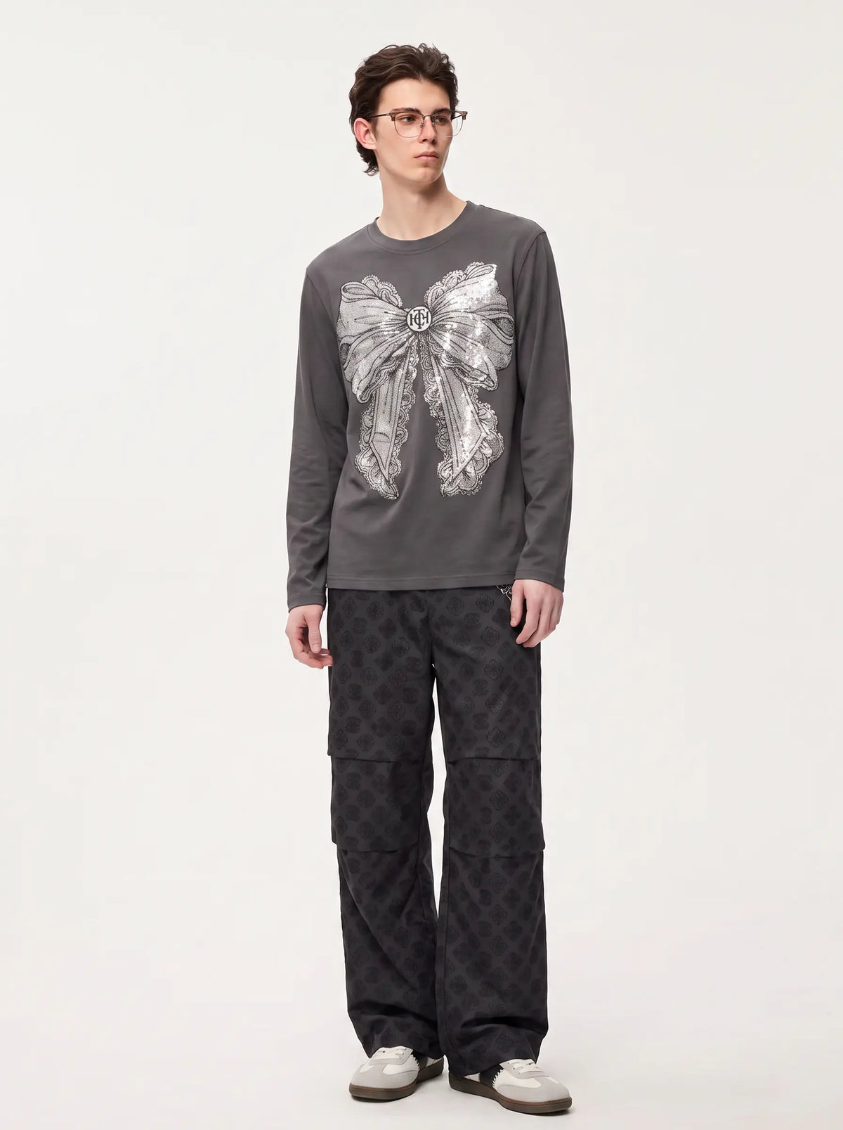 A man wearing a dark gray long-sleeve shirt with a large silver bow graphic on the front, paired with dark patterned cargo pants and white sneakers, standing against a plain light gray background. TCH Rhinestone Bow Graphic Fitted Long Sleeve Top