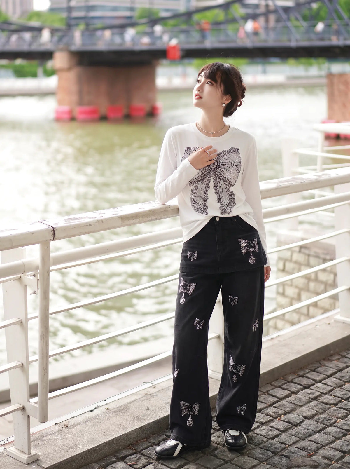 A woman stands outdoors by a river railing wearing a white long-sleeve top with a large gray butterfly graphic on the front and black wide-leg pants with matching butterfly prints. A bridge and water are visible in the background. TCH Rhinestone Bow Graphic Fitted Long Sleeve Top