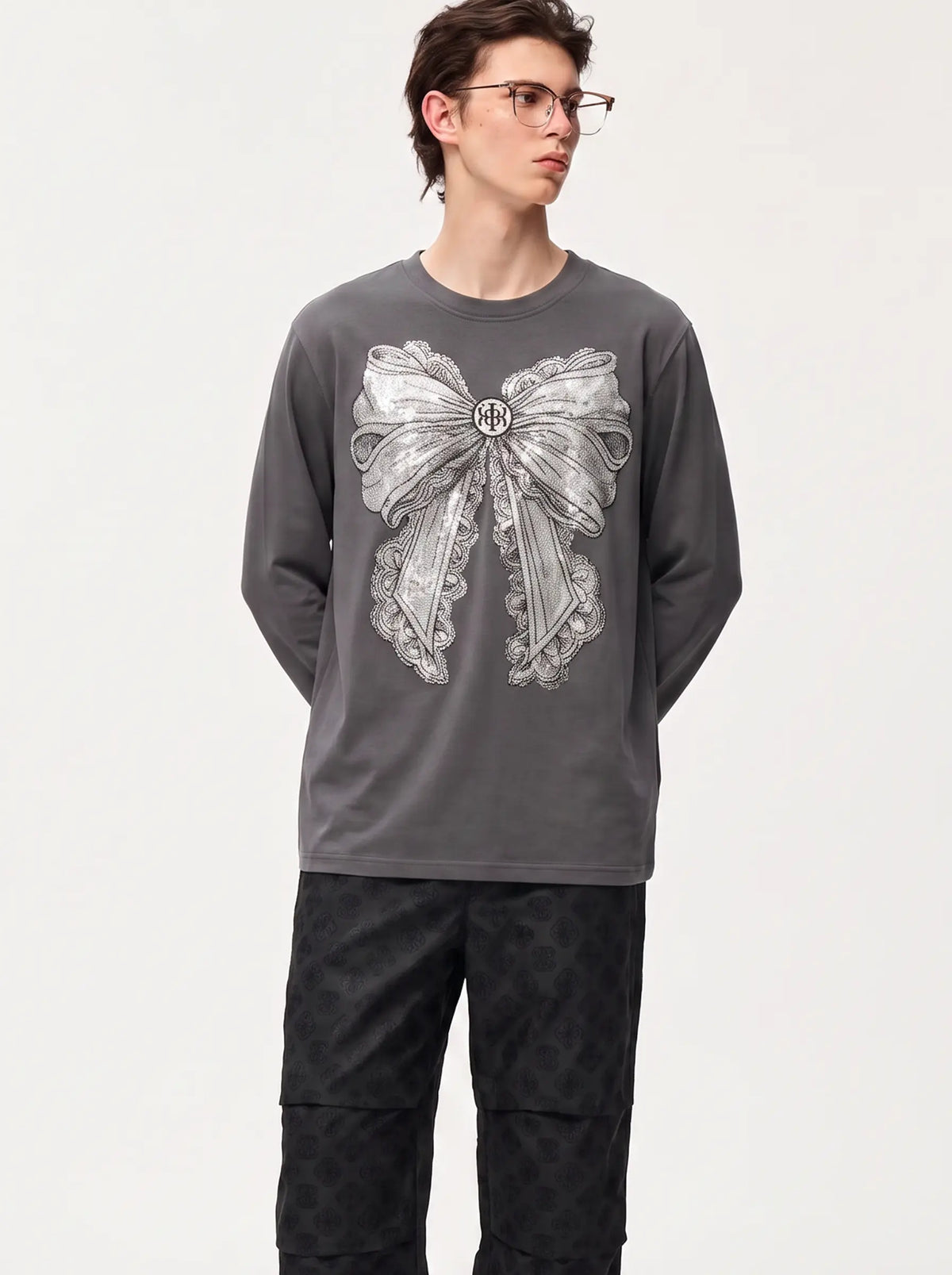 A model wearing a dark gray long-sleeve t-shirt with a large silver bow graphic on the front, paired with dark patterned pants, against a plain light gray background. TCH Rhinestone Bow Graphic Fitted Long Sleeve Top