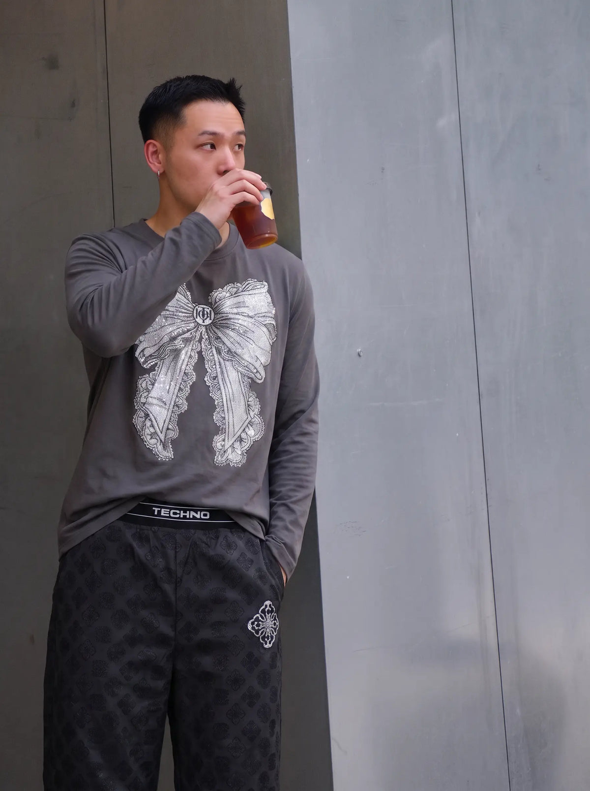 A man wearing a gray long-sleeve shirt with a large white bow graphic and black patterned pants, drinking from a brown bottle against a gray concrete wall. TCH Rhinestone Bow Graphic Fitted Long Sleeve Top